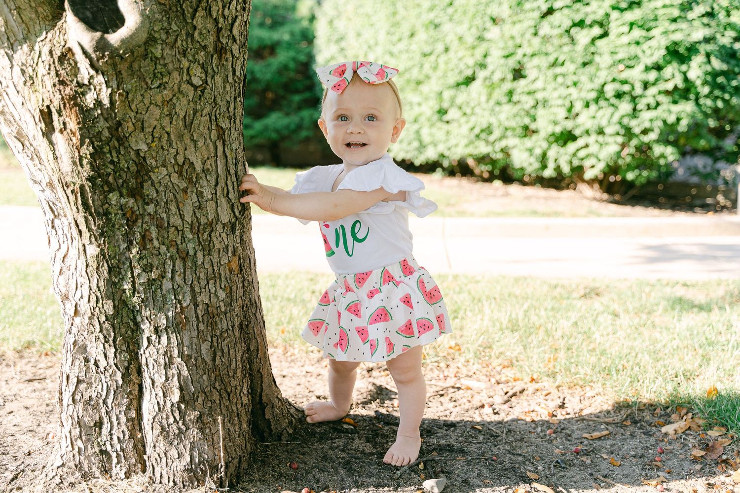 Baby wearing sage green ruffle outfit sits on white blanket outdoors in summer garden setting.