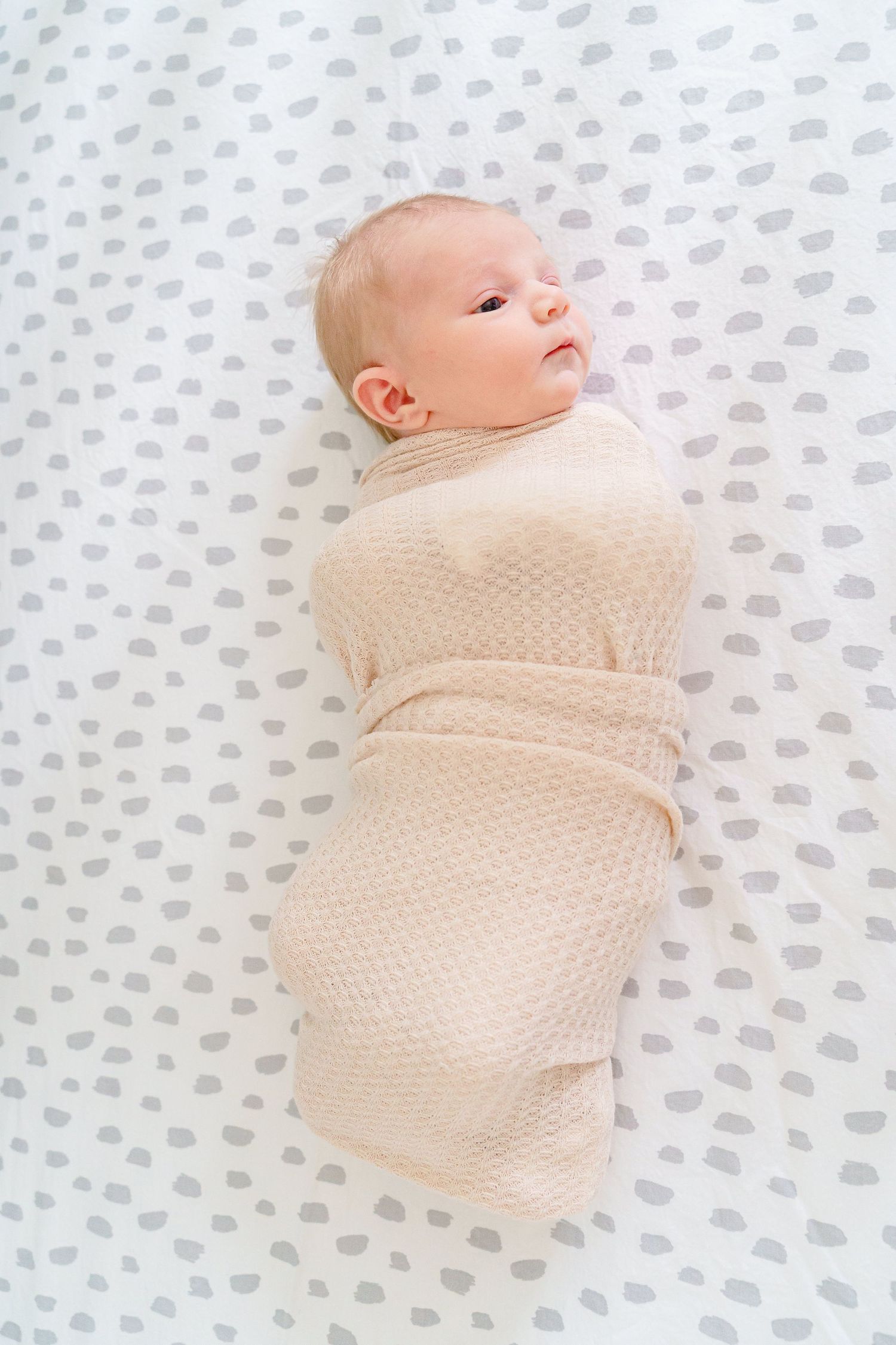 A newborn baby swaddled in a cream-colored blanket lies on a polka dot sheet looking upward.