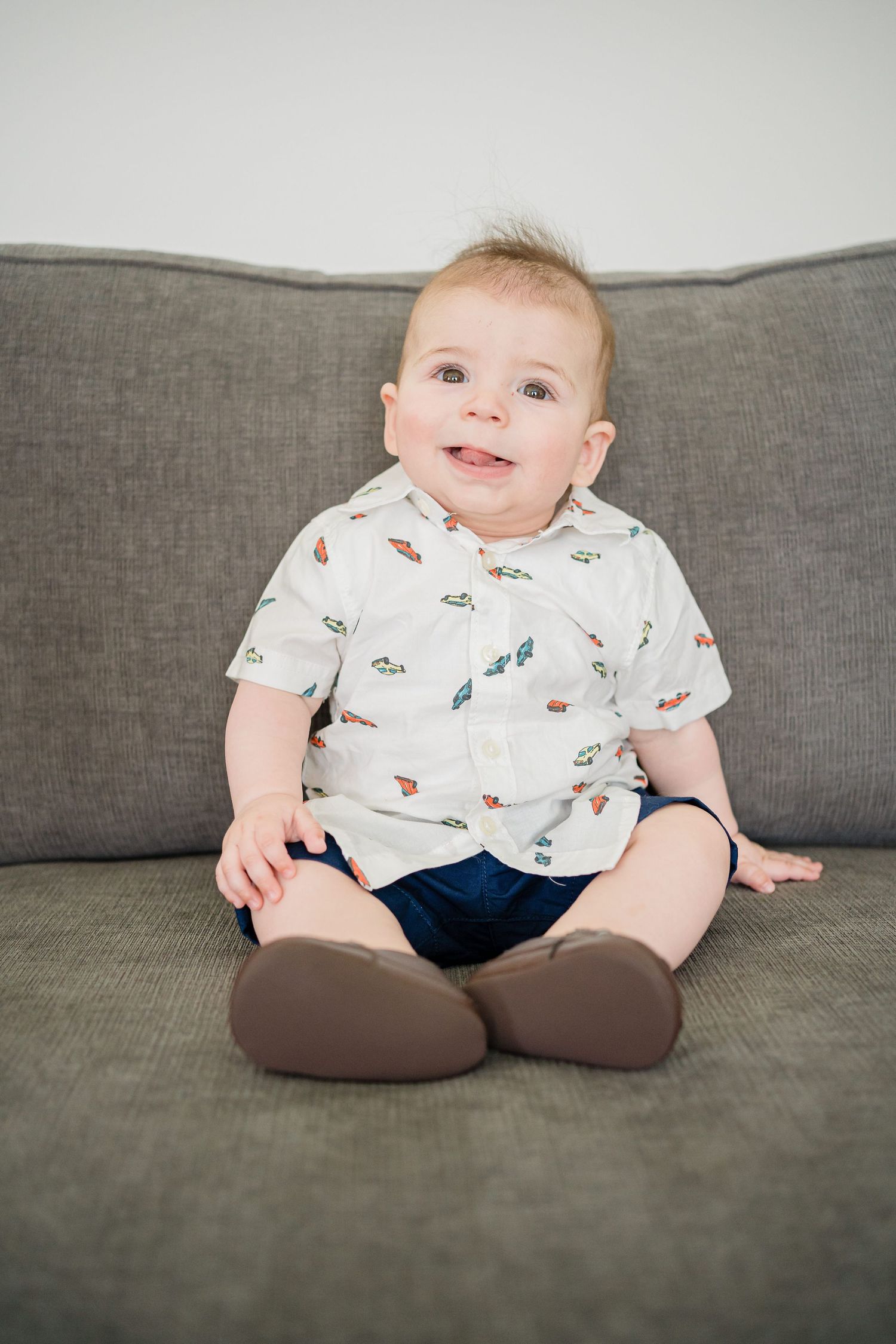 Baby wearing a patterned white shirt and navy shorts sits smiling on a gray couch.