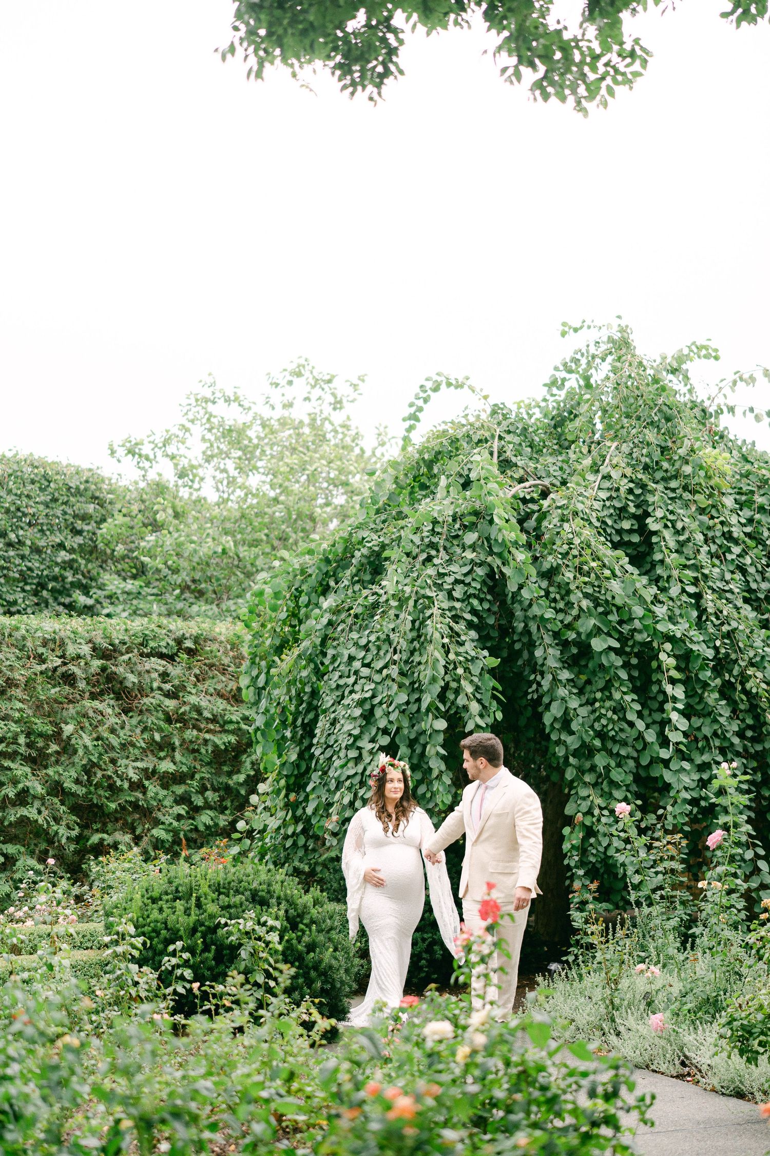 A couple walks through a lush garden path with cascading vines and blooming flowers on a sunny summer day.