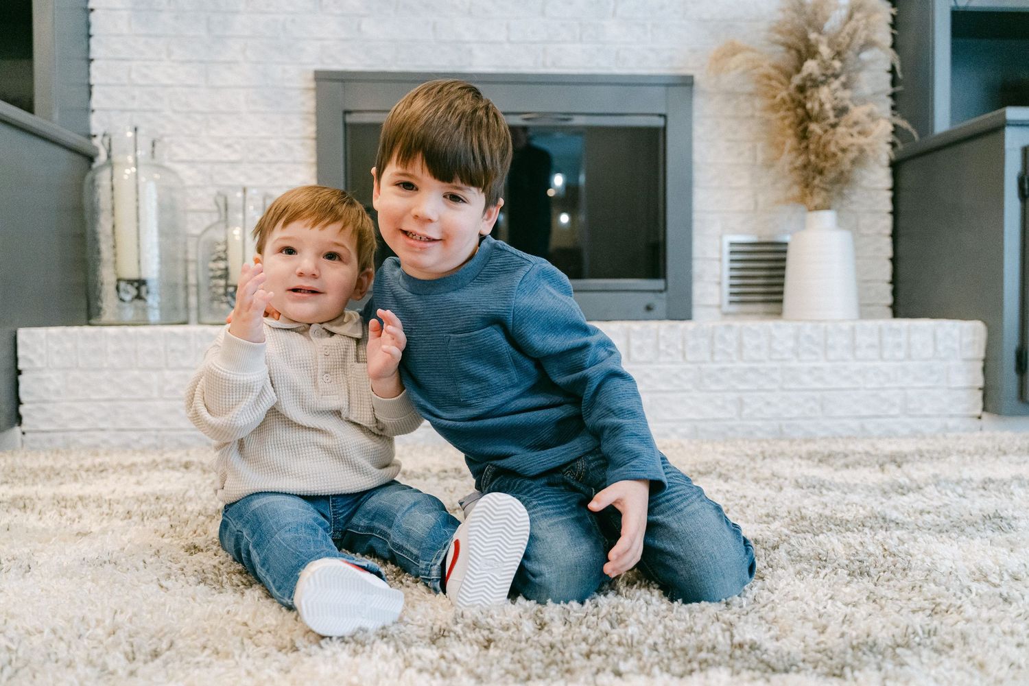 Two children sit together on a beige carpet in front of a white brick fireplace wearing casual clothing.