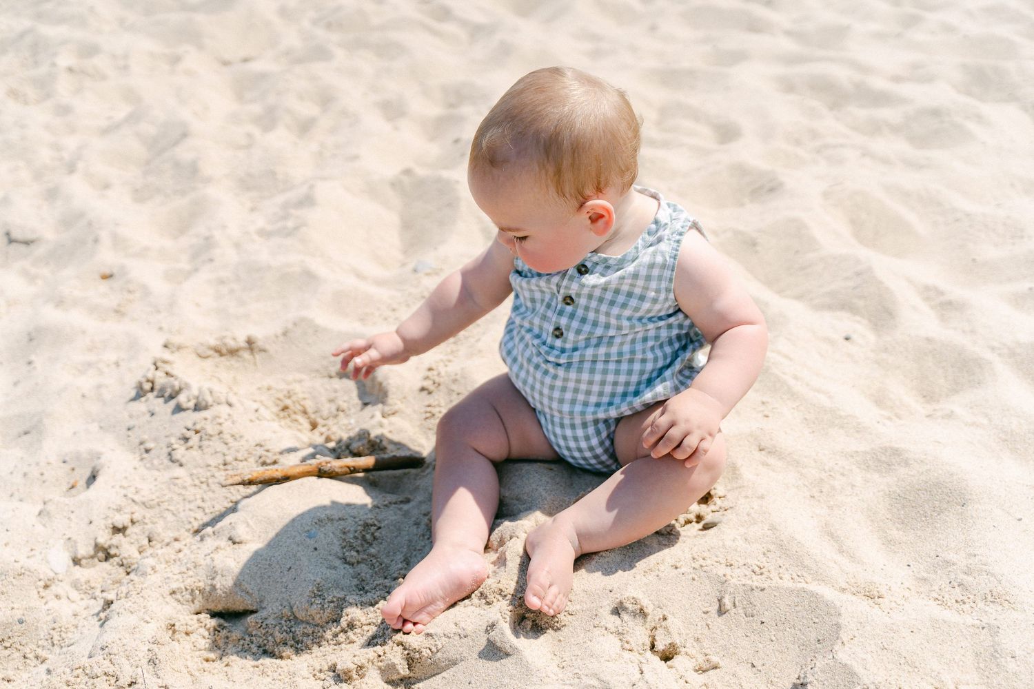 Baby in blue gingham outfit playing and exploring in the sand at the beach.
