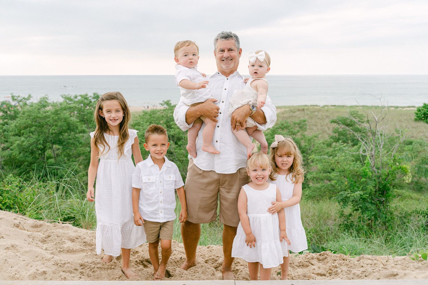 A group in coordinating white and neutral outfits poses together on a grassy hillside overlooking the ocean.