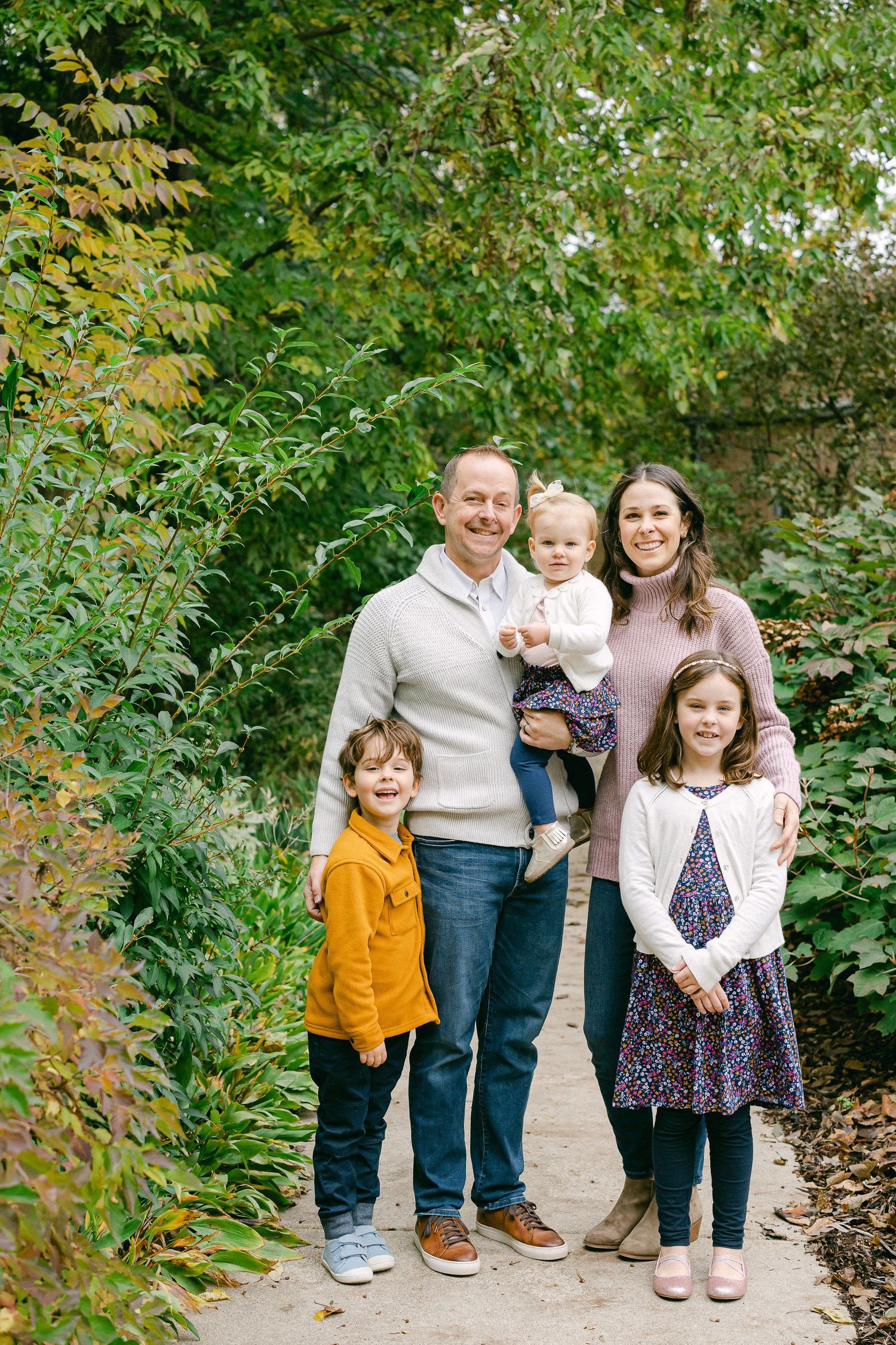 A family stands together on a garden path surrounded by green plants and autumn leaves.