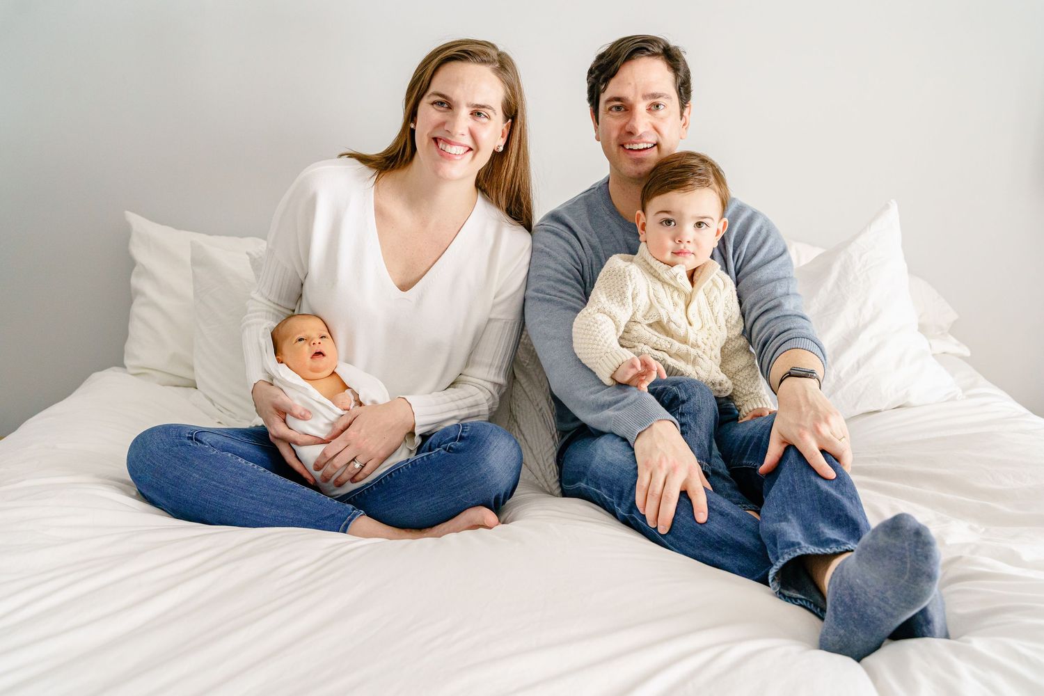 Family relaxing together on white bedding in casual clothing during lifestyle photoshoot.