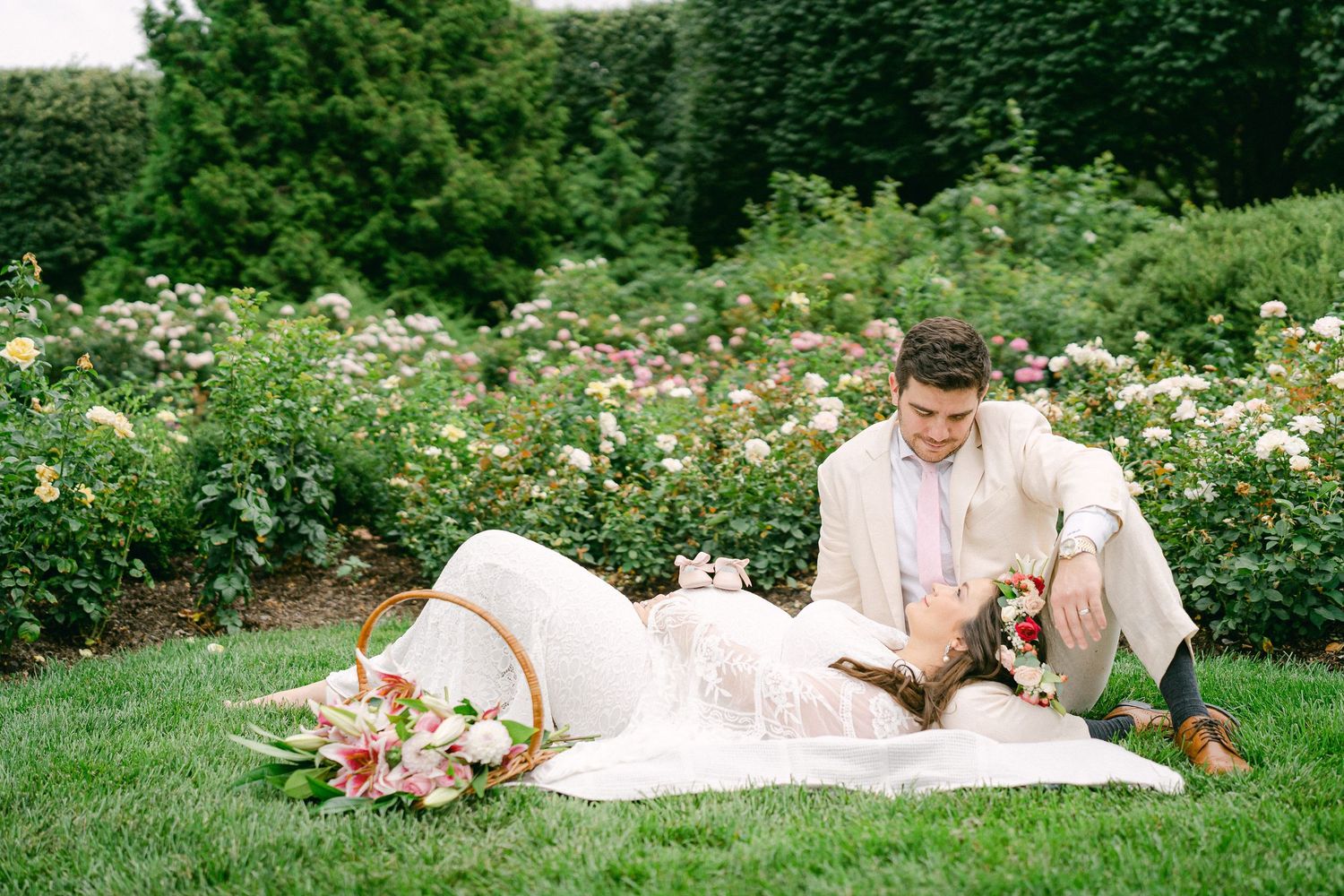 A romantic couple in wedding attire lies on grass in a garden surrounded by blooming summer flowers.