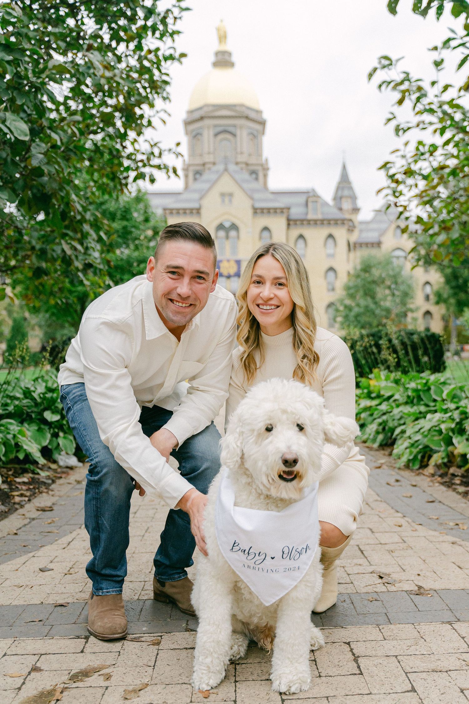 Couple poses with their fluffy white dog on stone pathway with grand historic building in background.