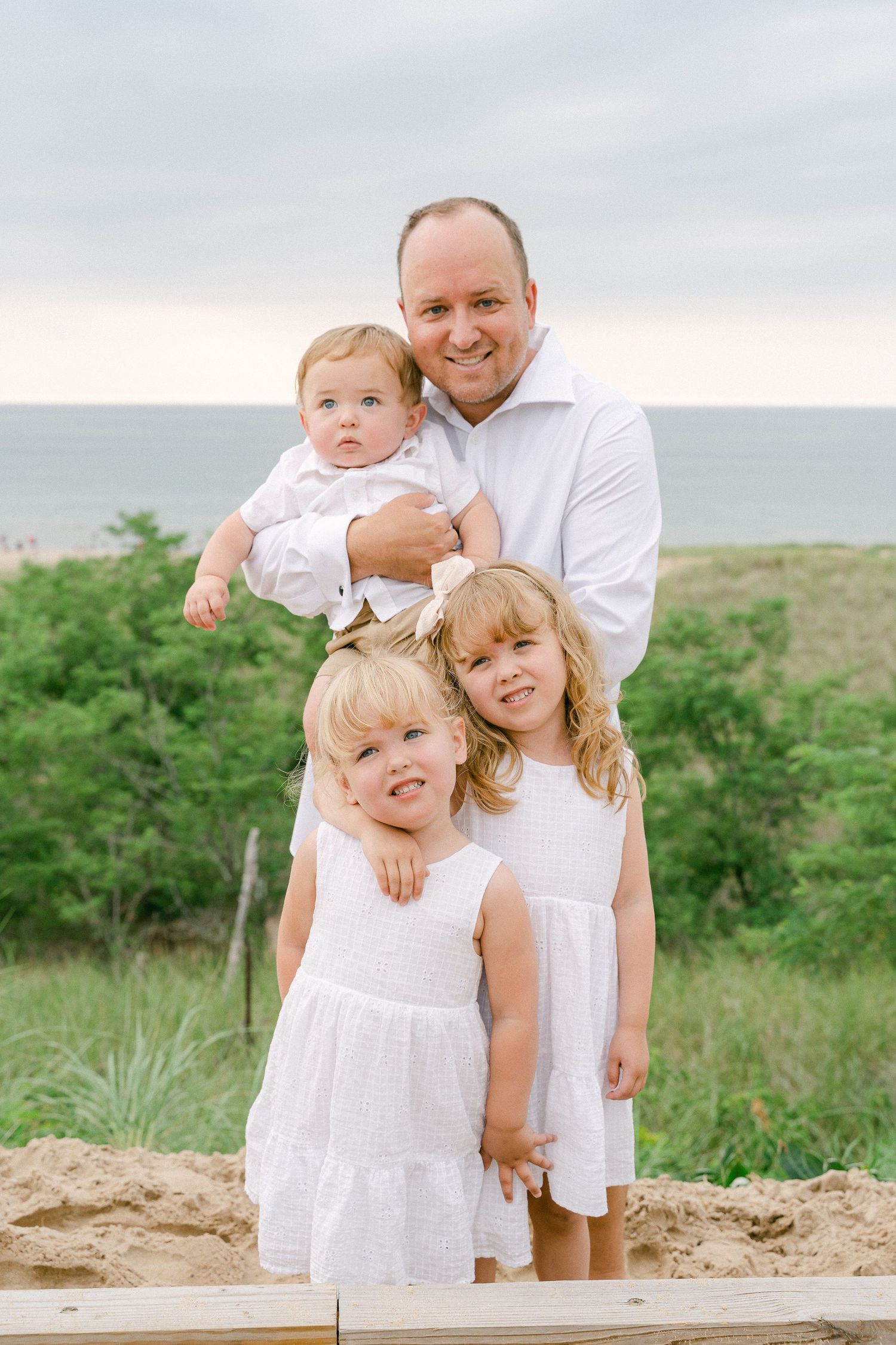 A family wearing white outfits poses together on a sandy beach with green foliage in the background during sunset.