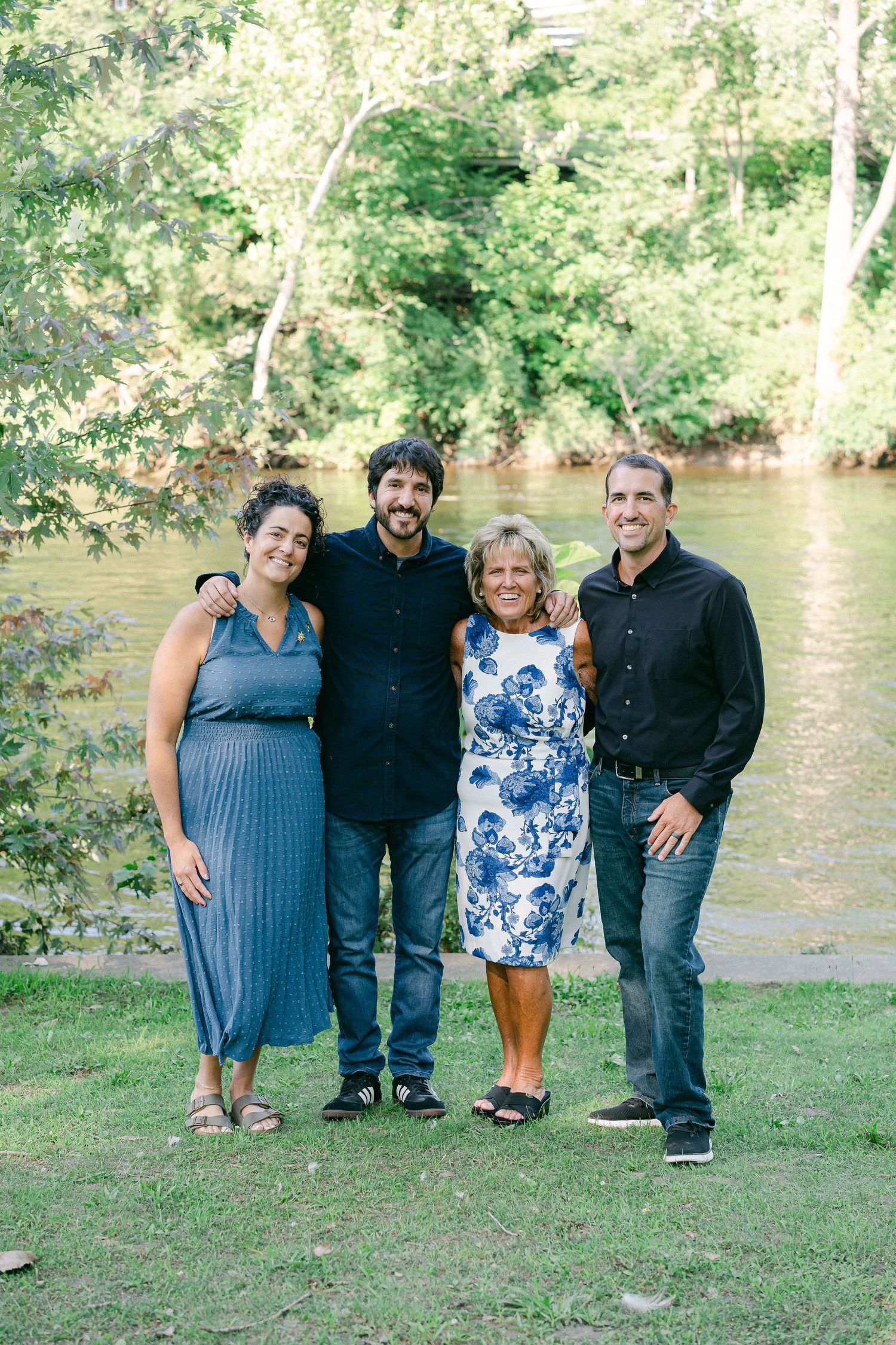 Four people stand together on grassy riverbank with trees and water in background on sunny day.