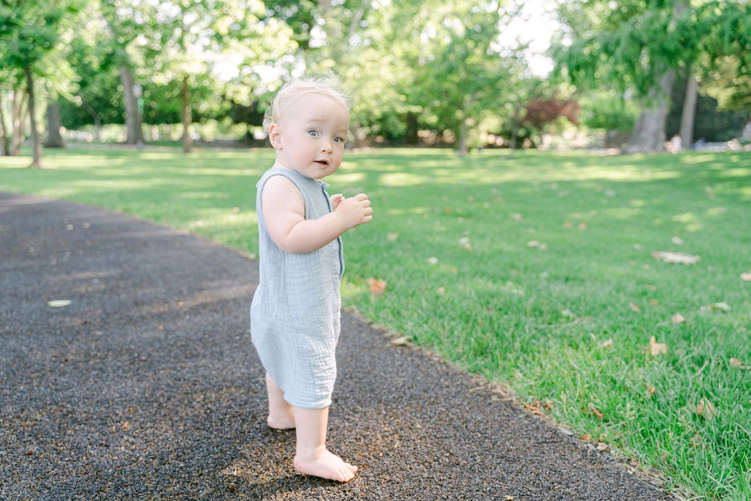 Small toddler in light blue overalls stands on a path surrounded by green grass in a sunny park.