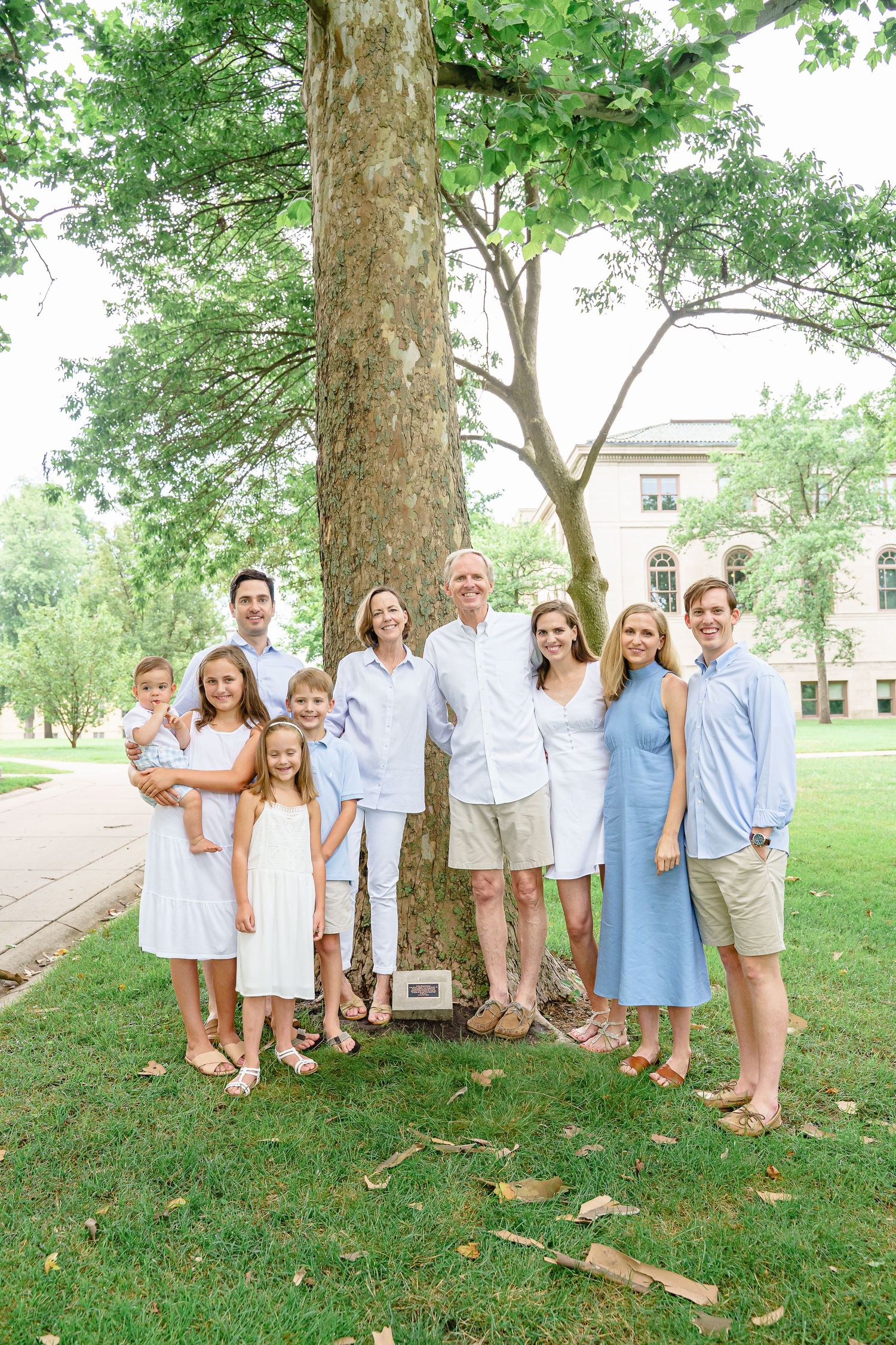 A family group poses together under large trees on a grassy lawn wearing coordinated blue and white summer outfits.