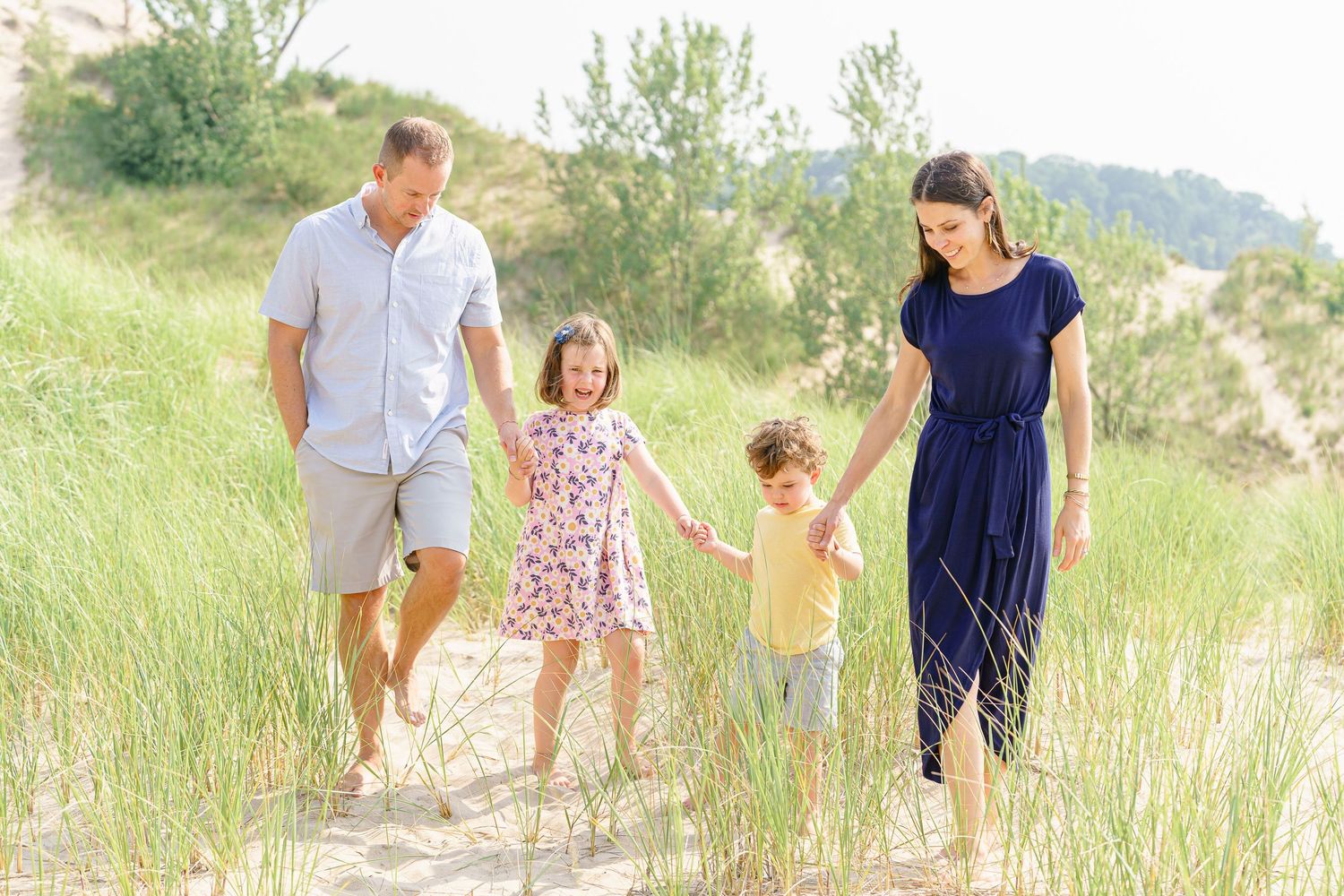 A family walks together through tall beach grass on a sunny summer day.