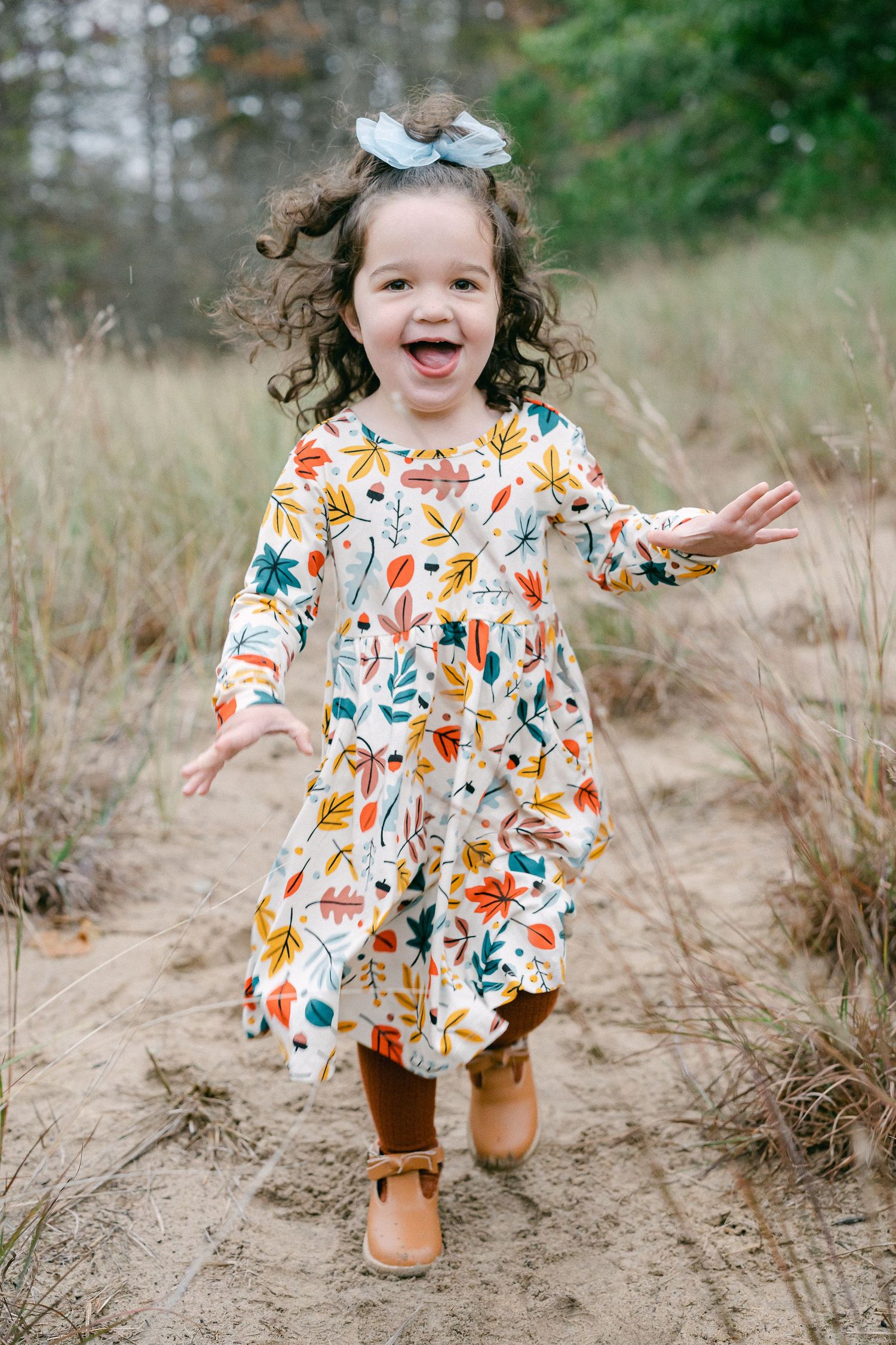 A joyful child in a colorful floral dress and brown boots runs along a dirt path through tall grass.