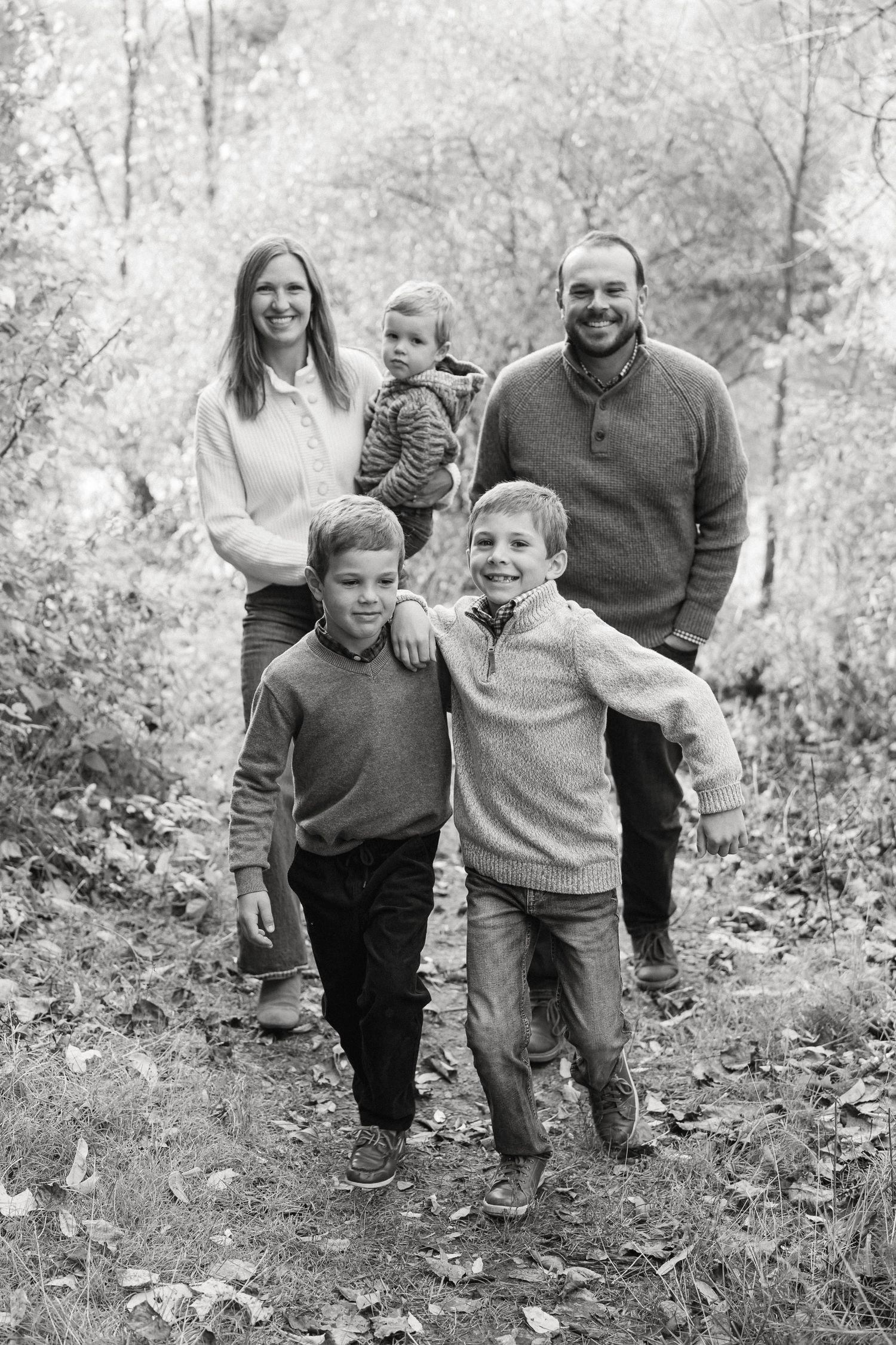 Black and white family portrait taken outdoors in a wooded setting during autumn with natural lighting.