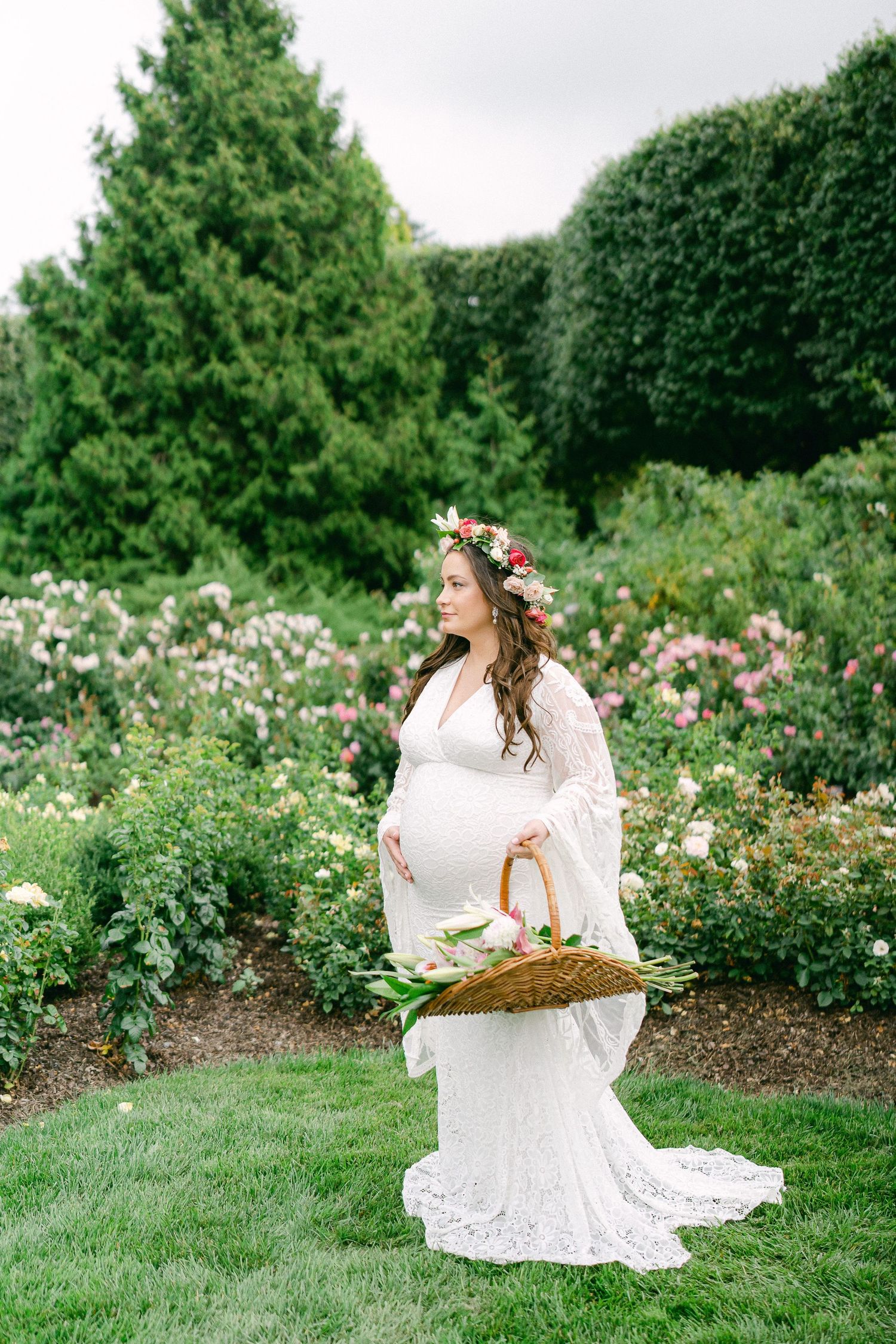 A woman in a flowing white dress sits gracefully in a blooming rose garden on a summer day.