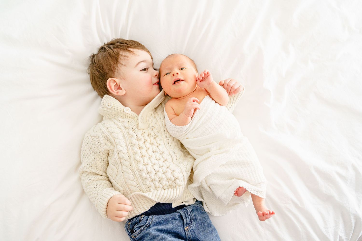 Two siblings sharing sweet moments on white bedding during indoor photoshoot.
