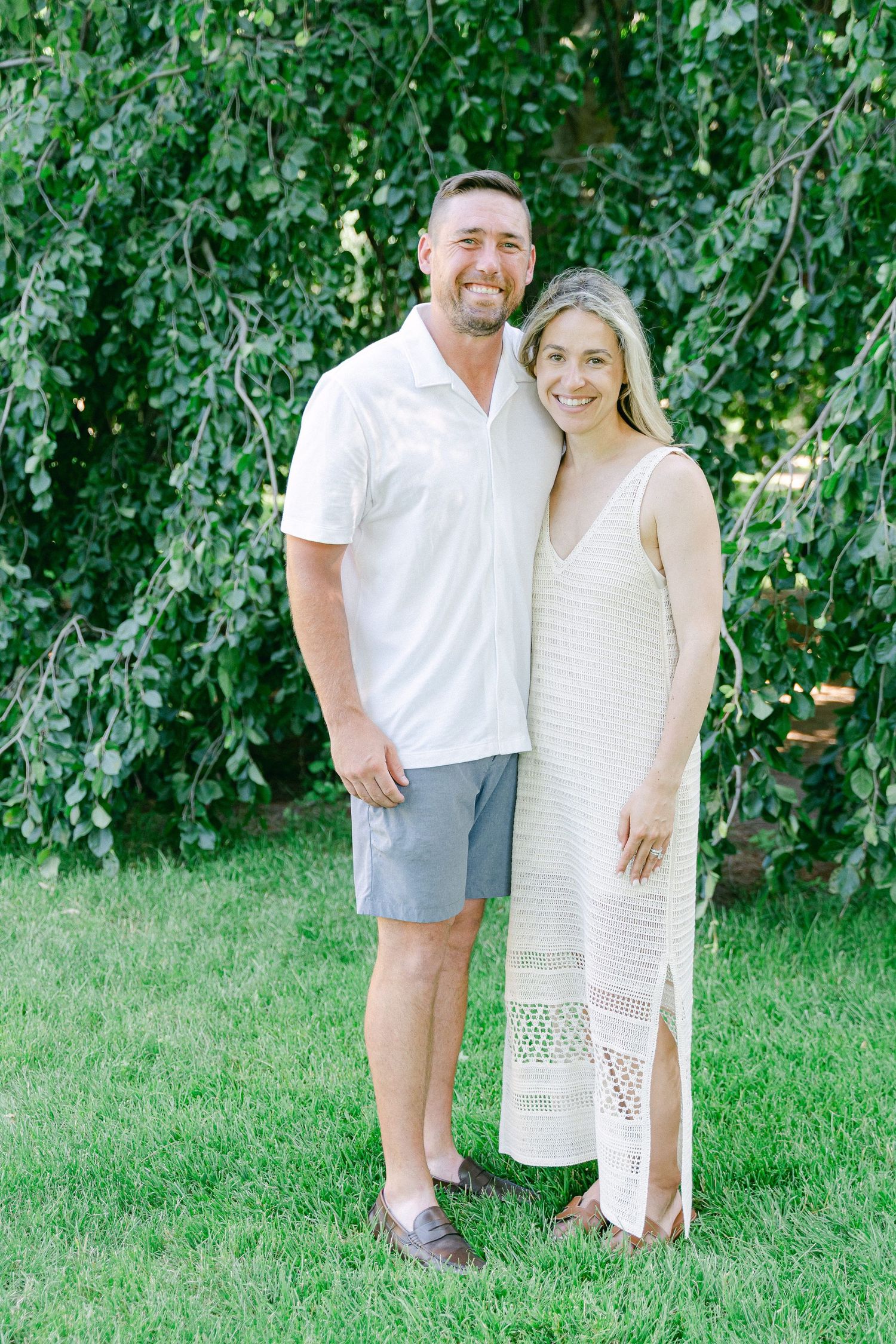 A couple in white outfits poses together on a green lawn with leafy trees in the background.