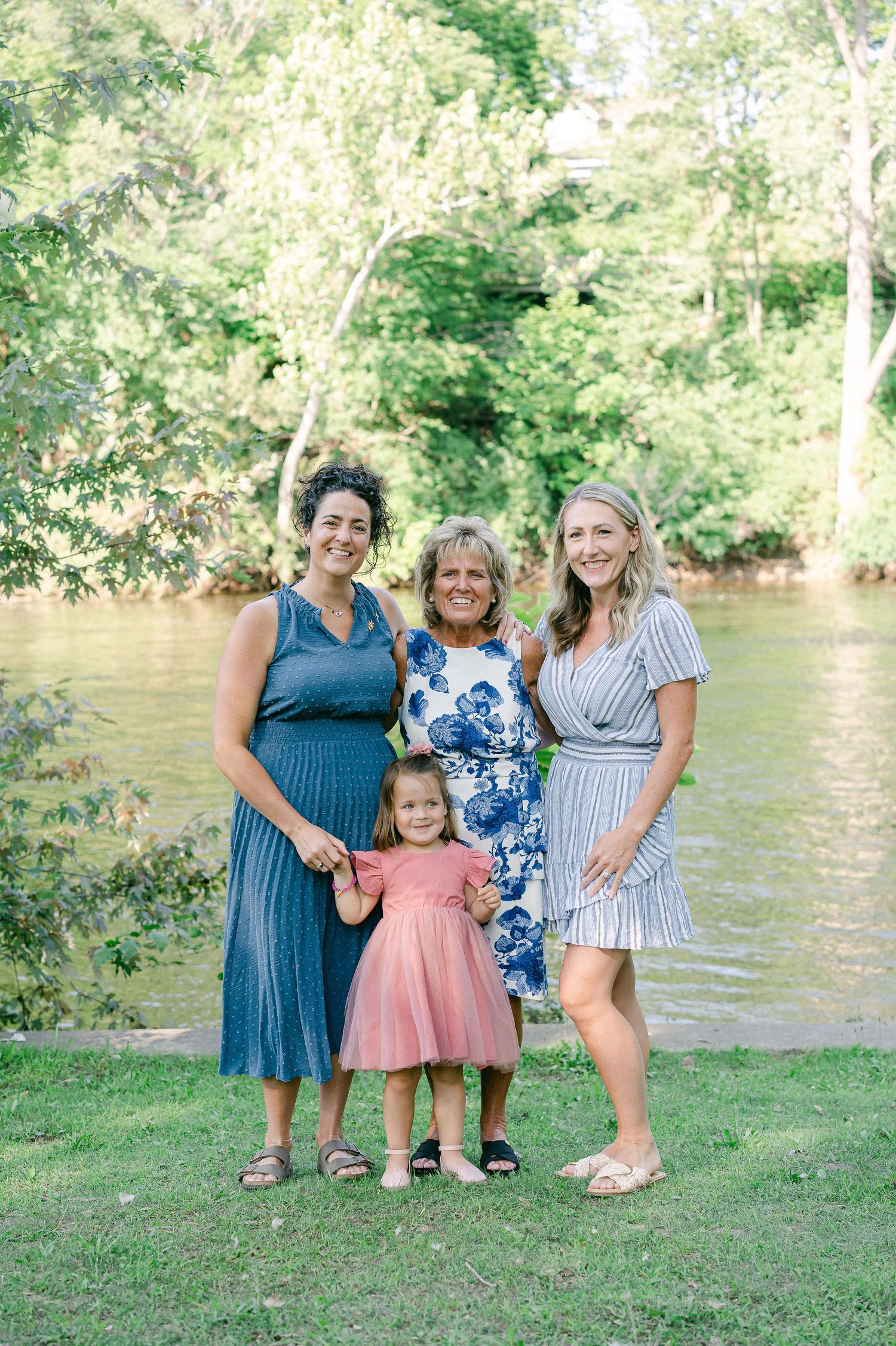 A group poses together by a river on a sunny day, wearing summer dresses in blue, floral and pink.