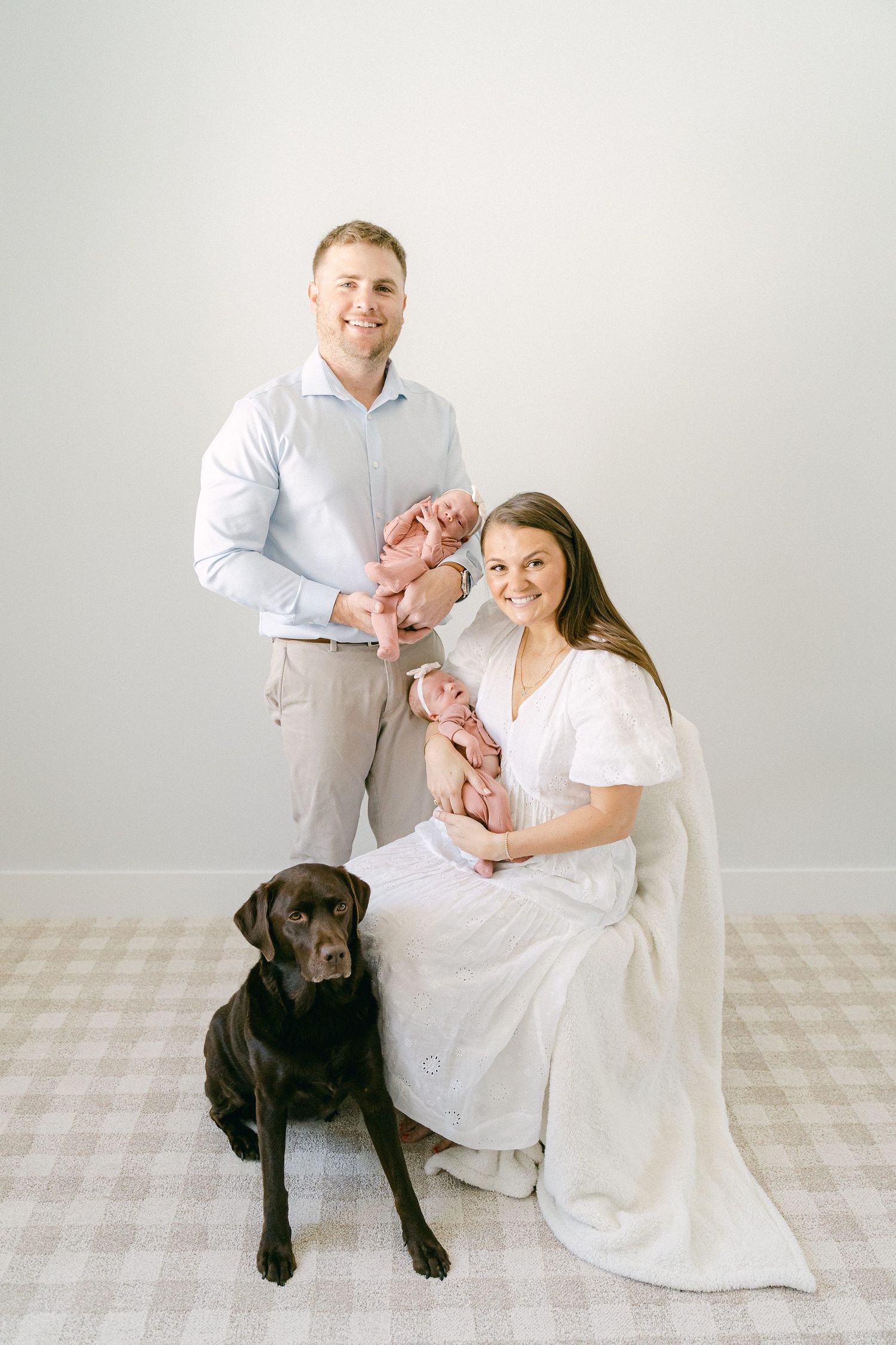 A family in white outfits sits together with their black Labrador dog and newborn baby in a minimalist studio setting.