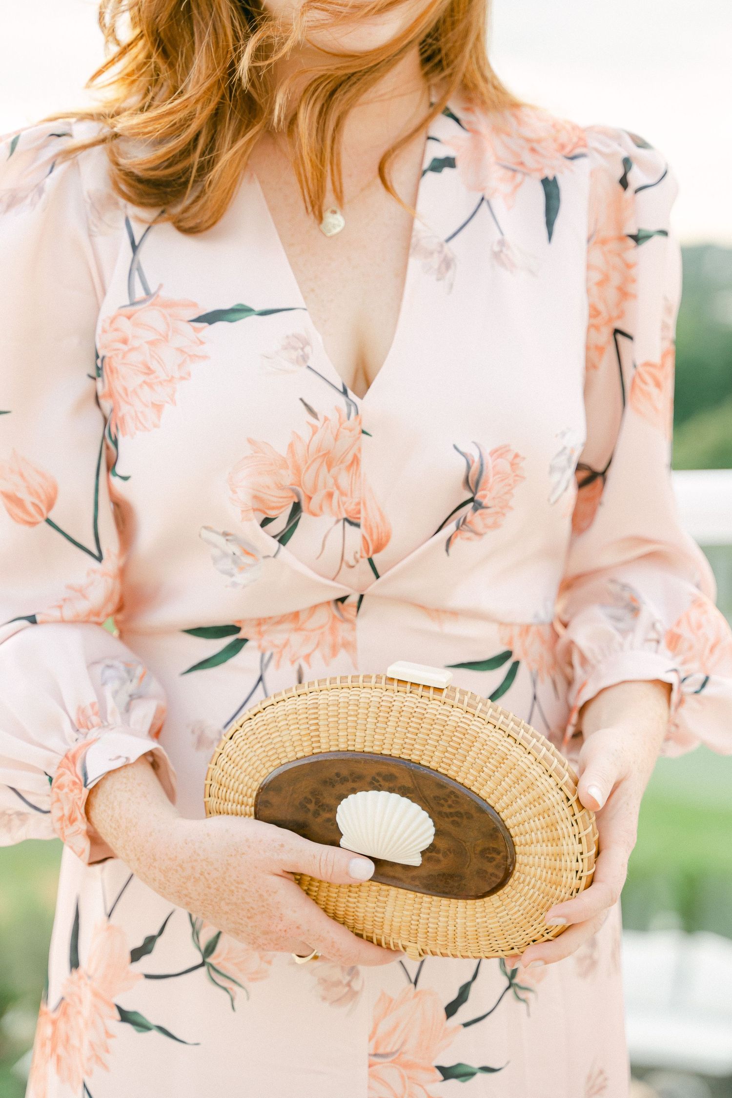 Woman in floral blush dress holding a woven rattan clutch with shell detail in Cape Cod