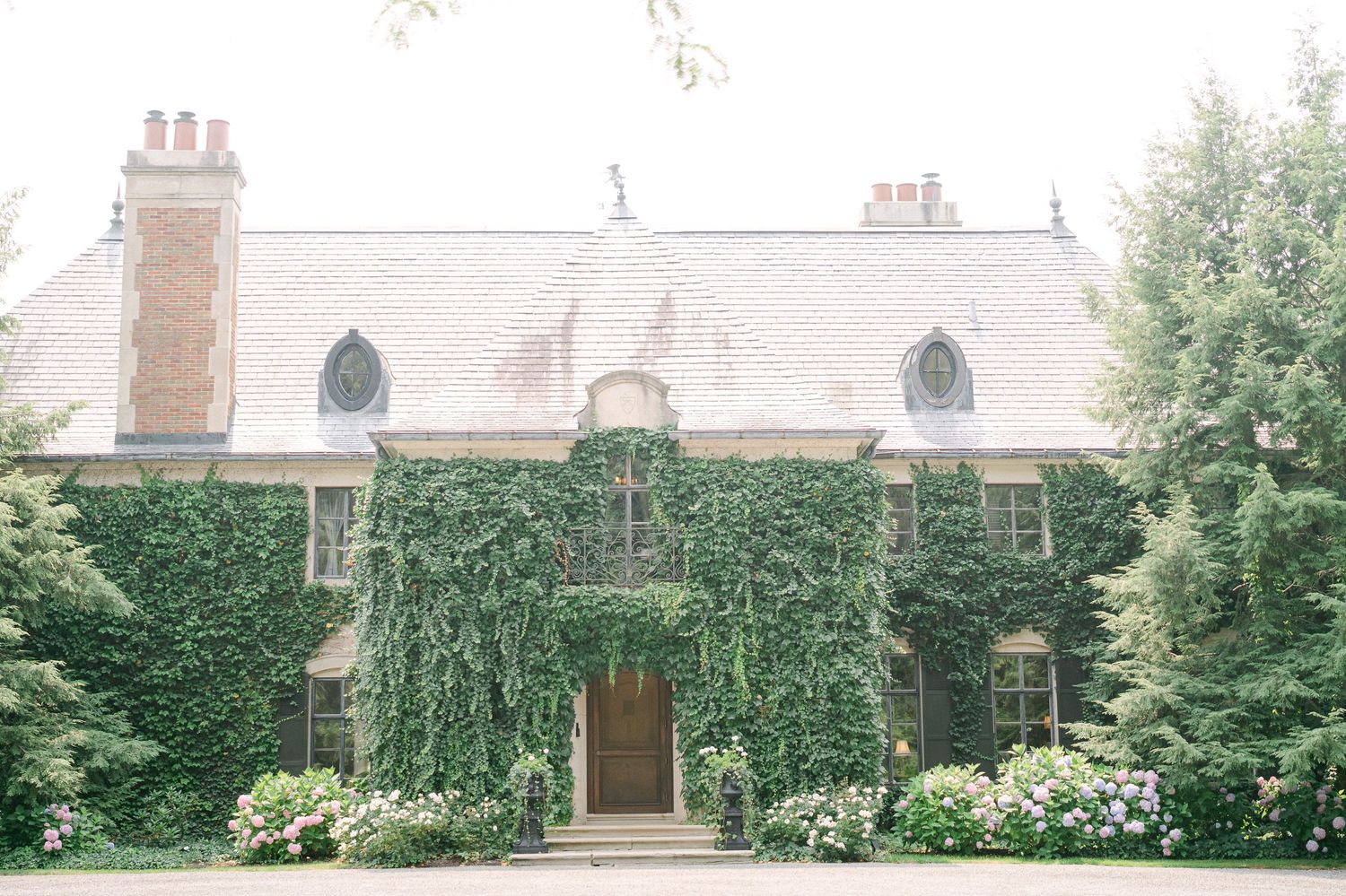 A panoramic view of a historic stone building with ivy-covered walls and manicured gardens in summer.