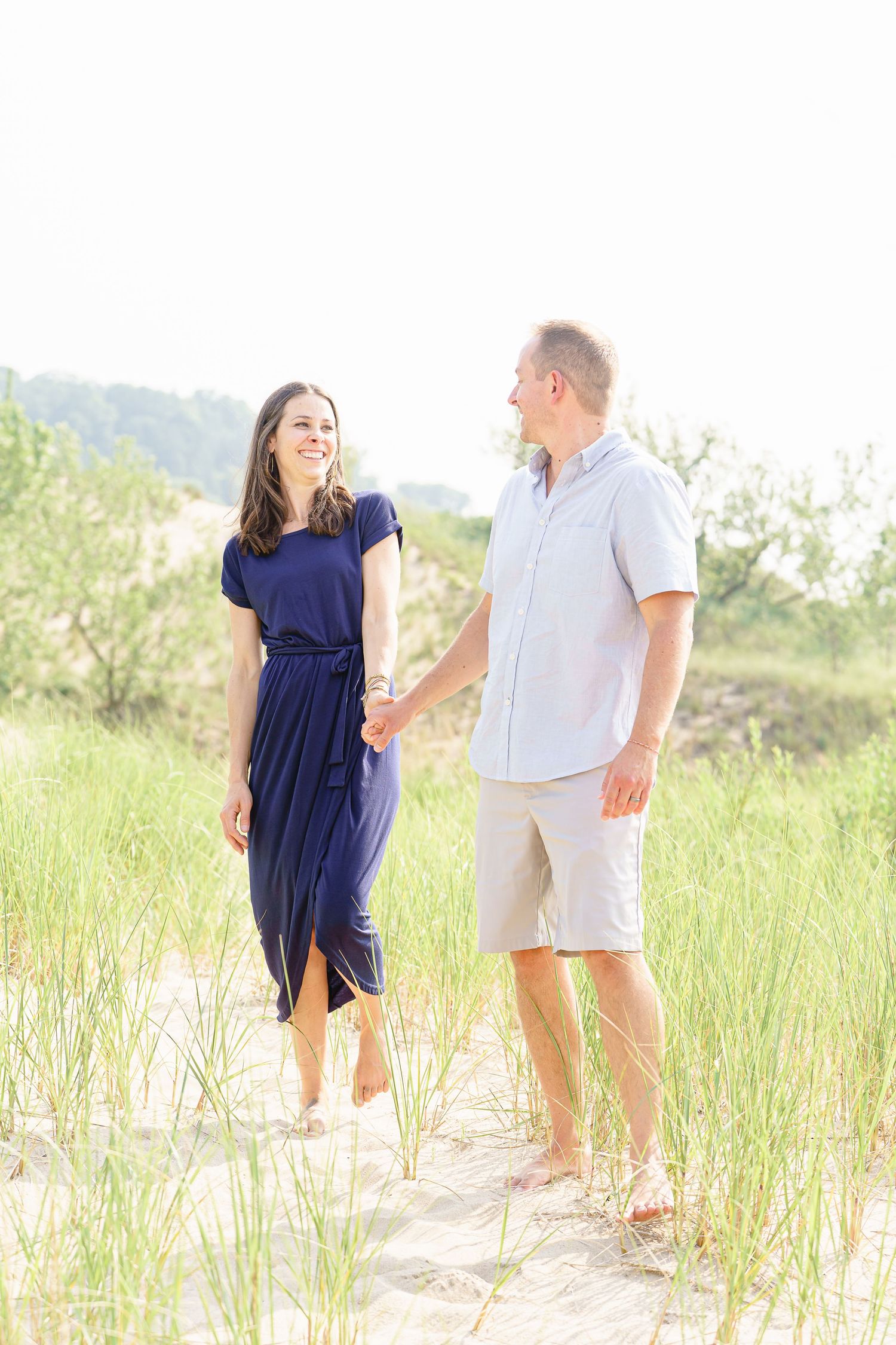 People stroll along a sandy beach path with tall grass and dunes in soft sunlight.