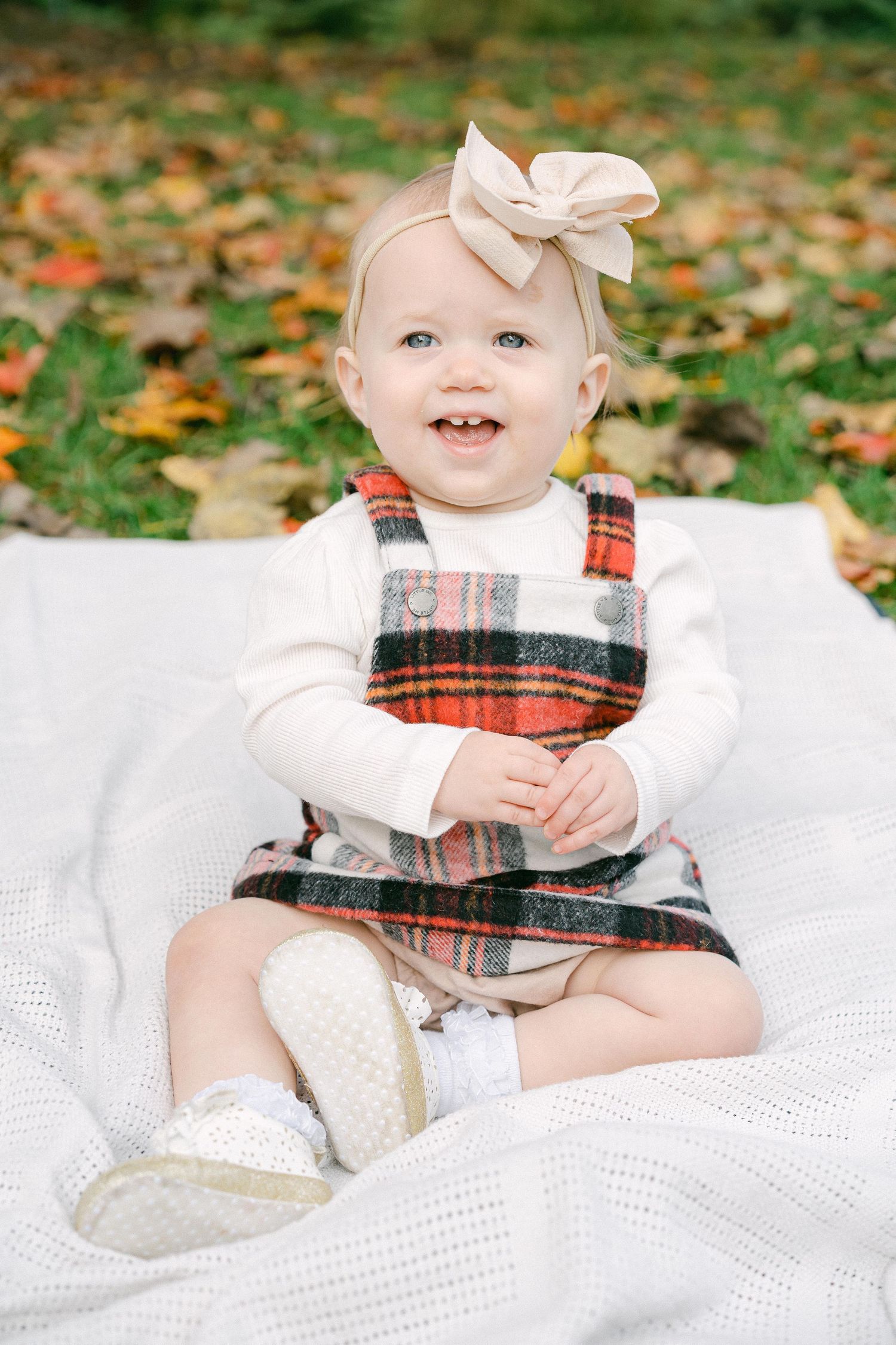 Baby wearing plaid jumper dress sits on white blanket surrounded by autumn leaves on grass.