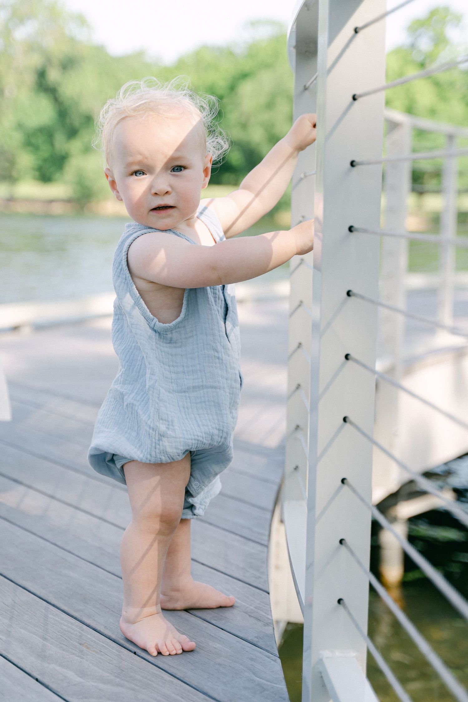 A small child in a blue romper explores a white railing on a dock by the water on a sunny day.