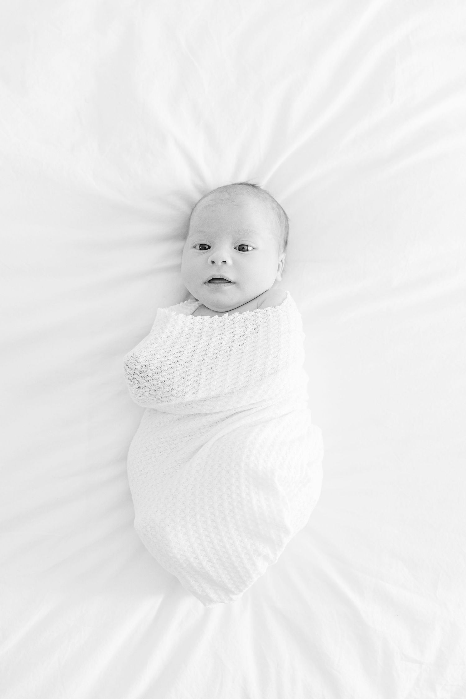Newborn baby wearing white swaddled on white bedding in minimalist style portrait.