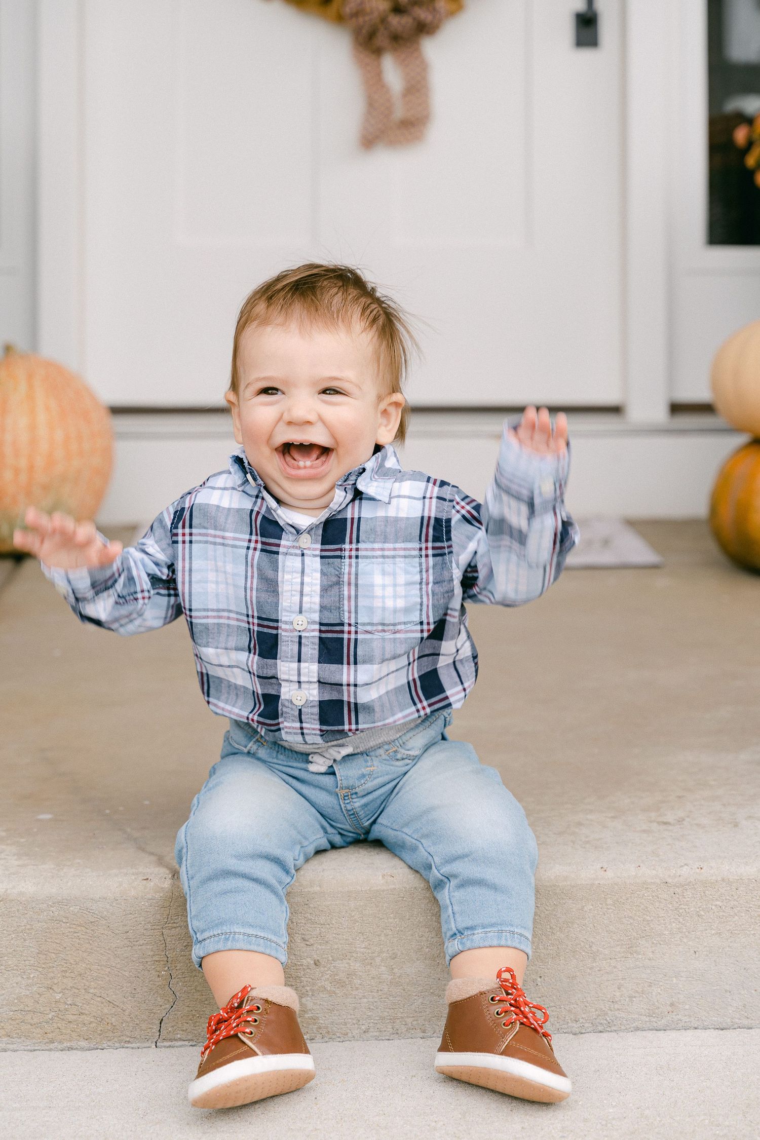 Baby in plaid shirt and jeans sits on porch with pumpkin decor, showing excited expressions.