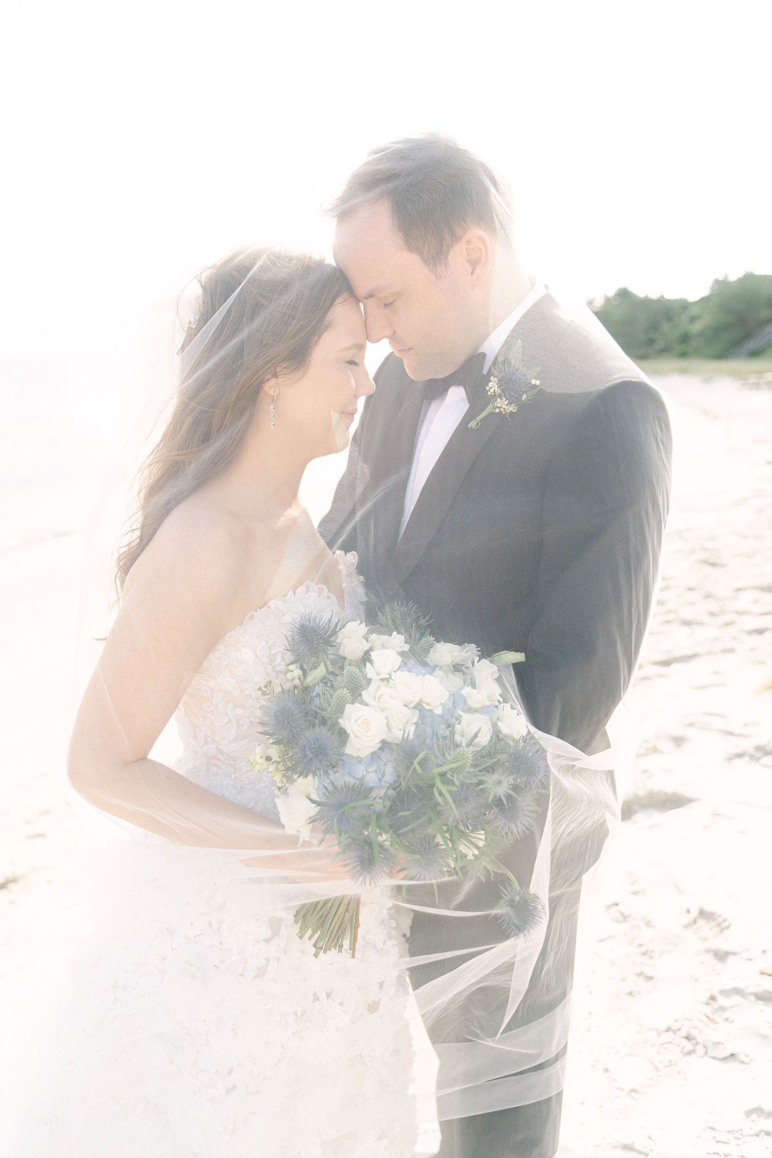Bride and groom sharing a romantic beach moment, her veil flowing, holding a blue and white bouquet in soft sunlight in Cape Cod