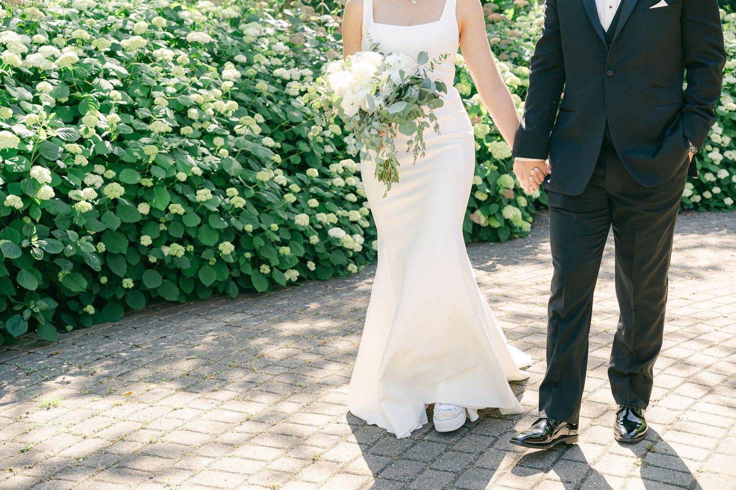 Bride in white mermaid gown holding bouquet walks with groom in black suit on brick path beside lush green hedges in Valparaiso, IN