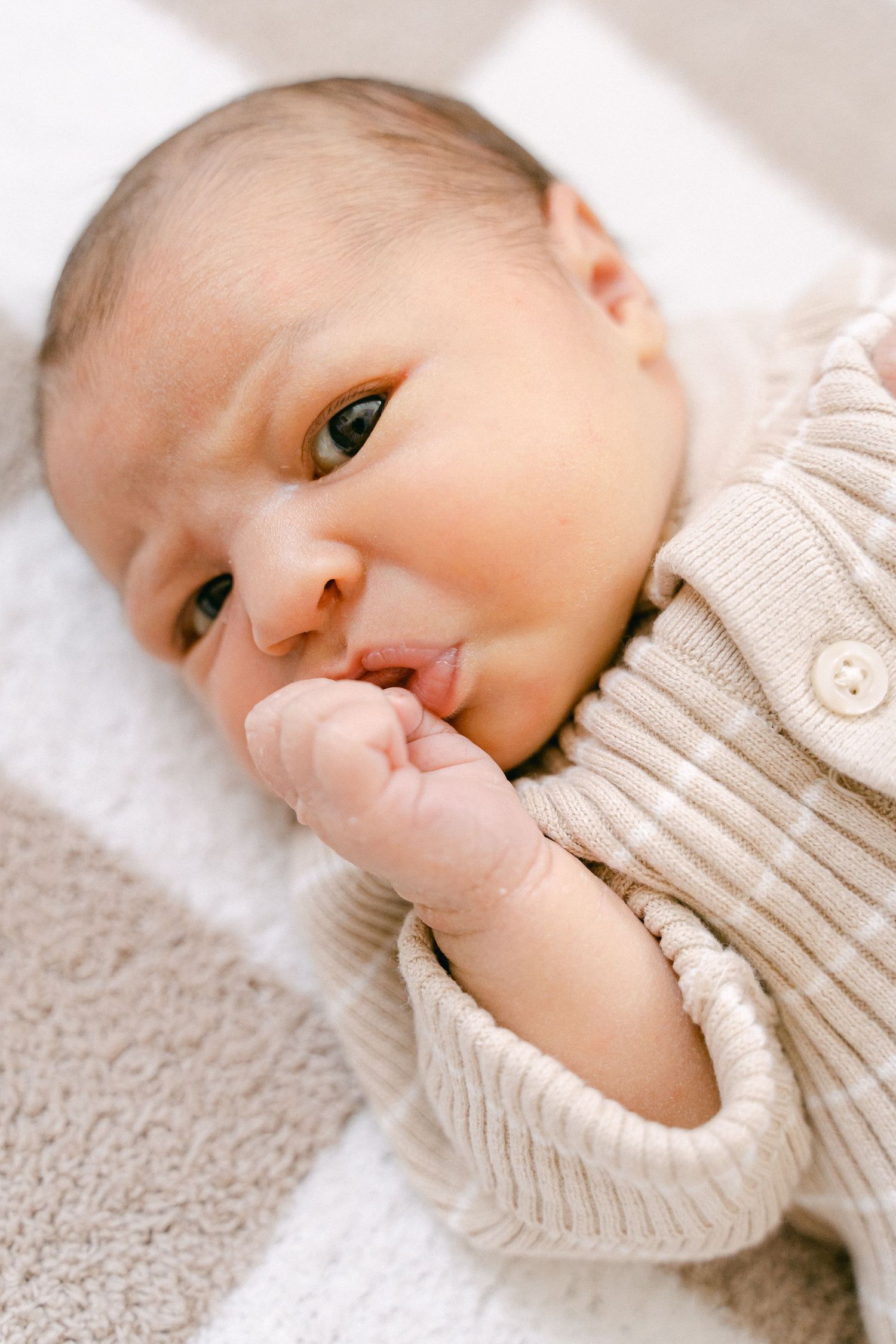 A sweet newborn baby in a cream knit sweater sucking their thumb while resting on soft fabric.