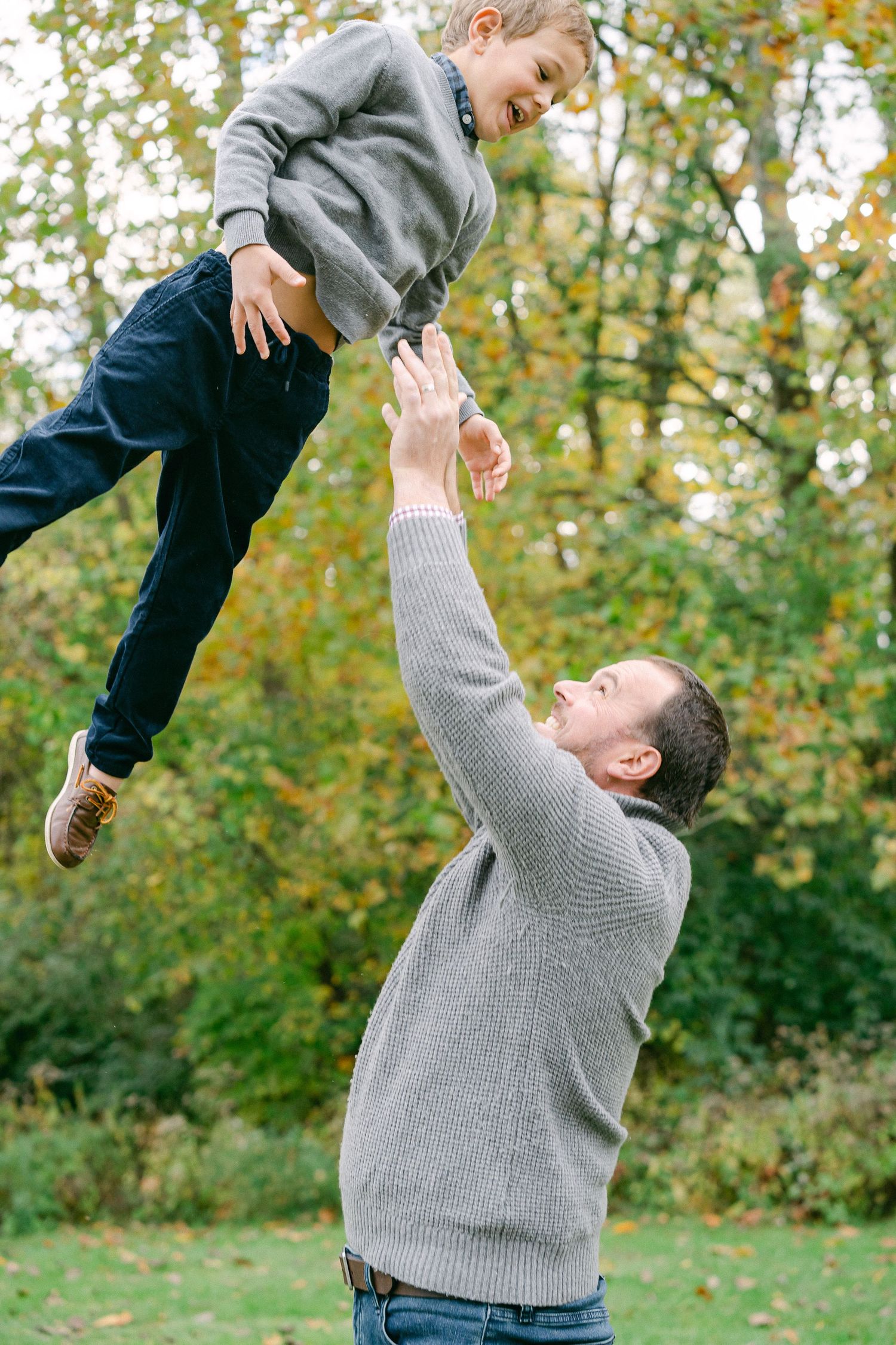 A joyful moment captured in autumn as a person in a grey sweater lifts a small child up against a backdrop of fall foliage.