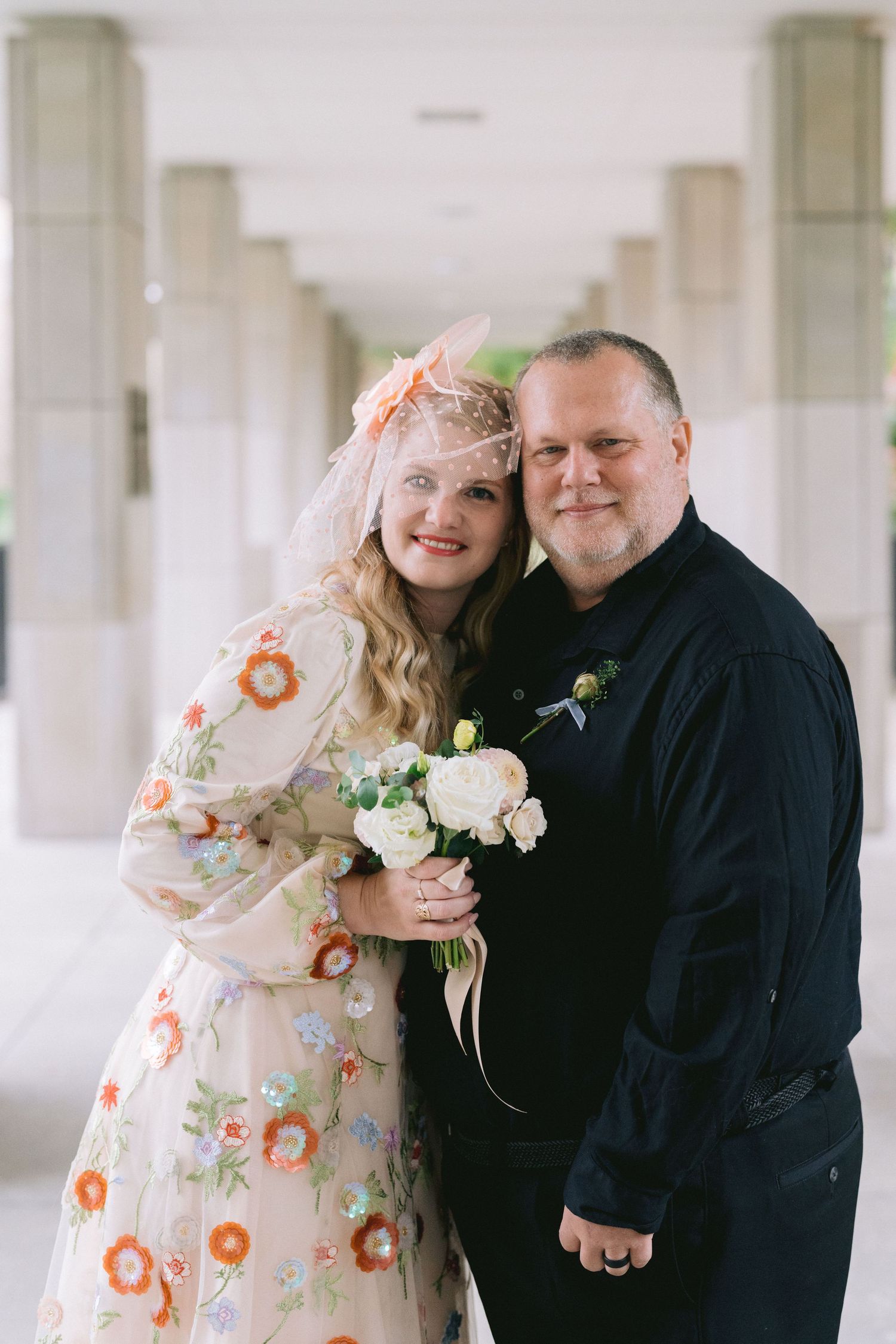 A couple shares romantic moments in a white hallway, with the bride wearing a floral dress and groom in navy.