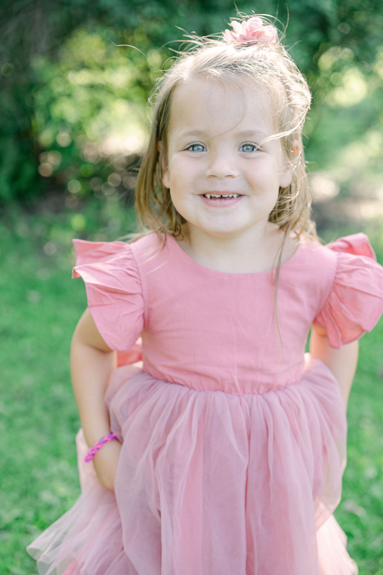 A young child in a pink ruffled dress smiles outdoors against a green background.