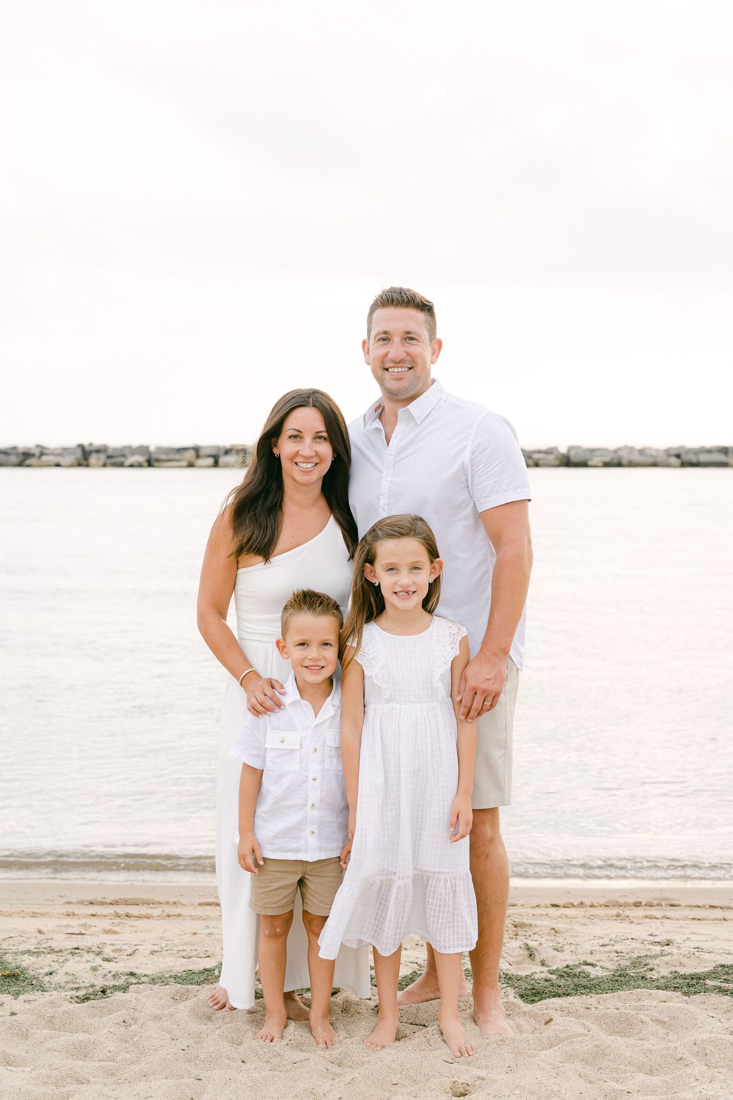 Family in coordinated white outfits poses together on beach with water view.