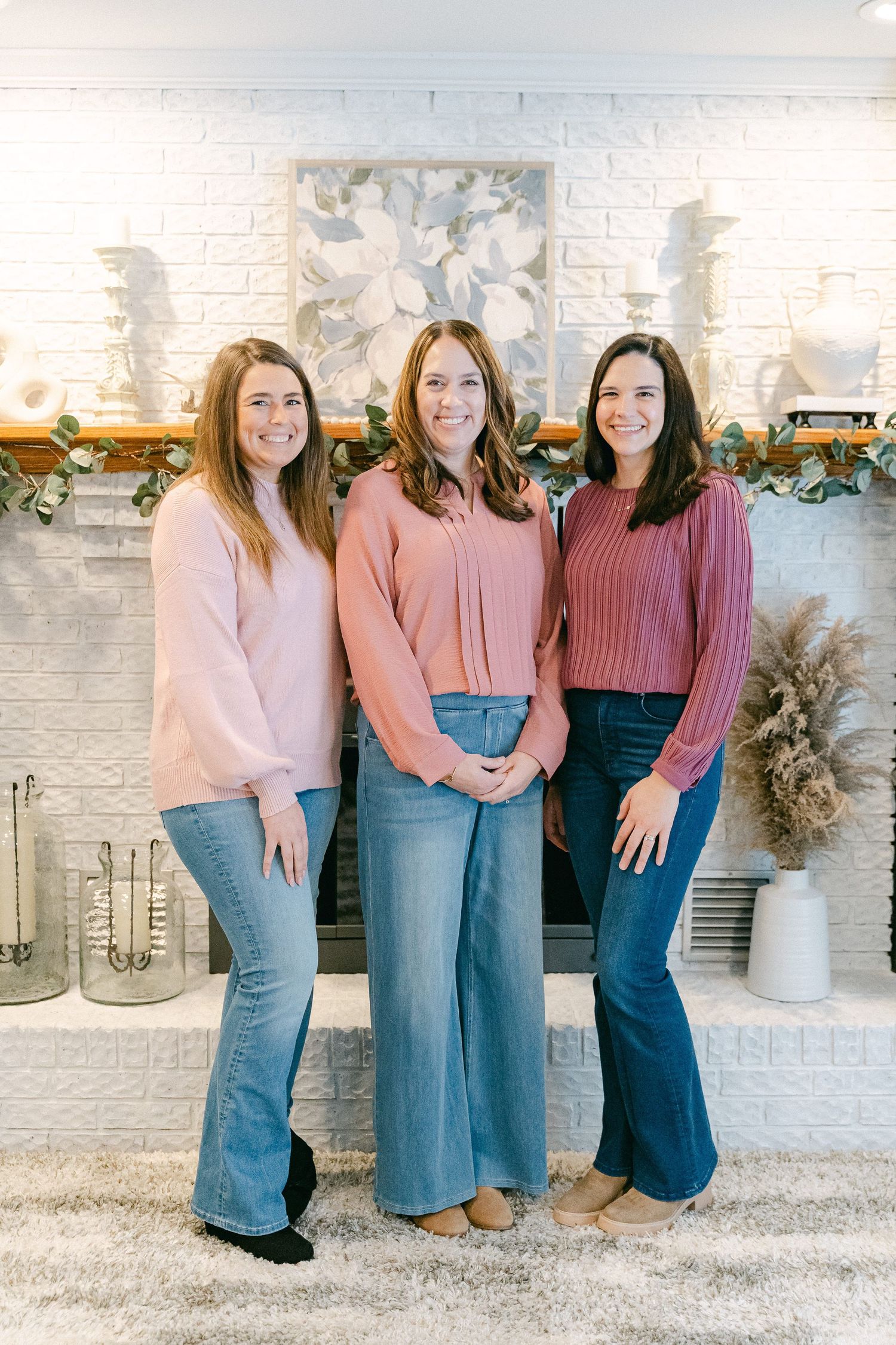 Three women in matching pink sweaters and denim skirts pose against a white brick wall with decorative accents.