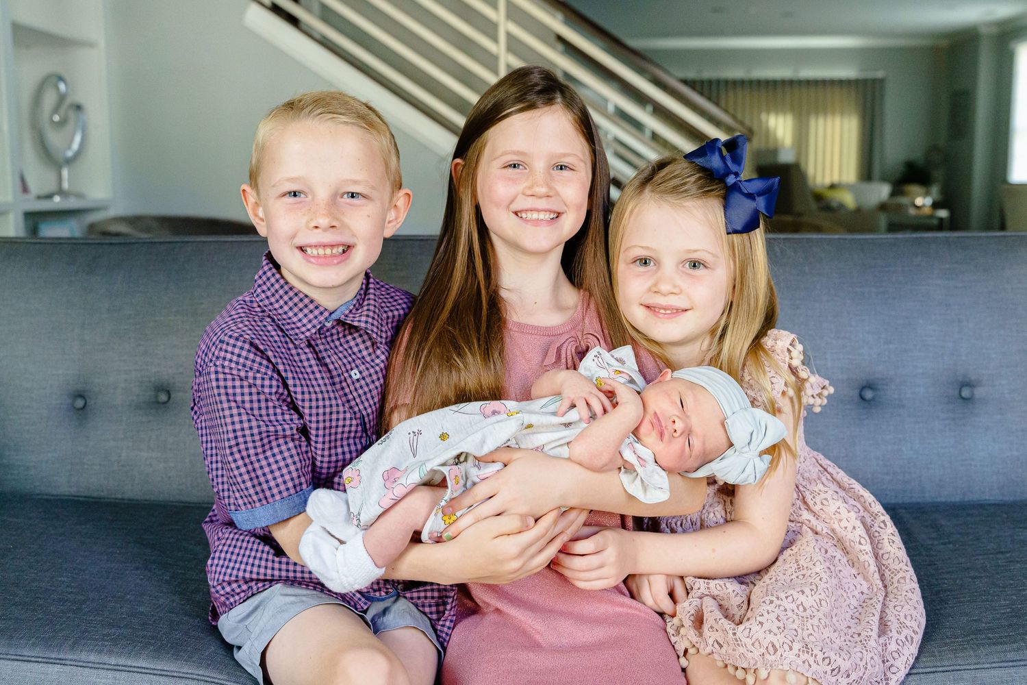 Three siblings sit together on a gray couch while holding their newborn baby sibling in a pink blanket.