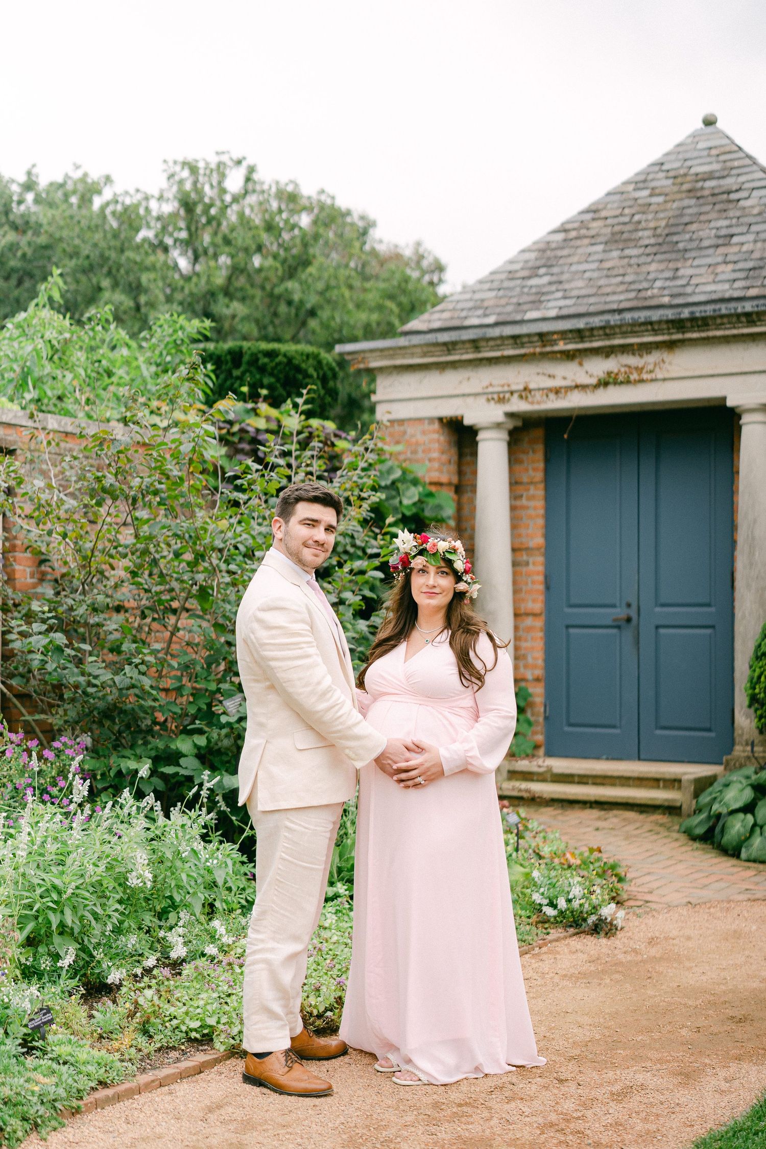A couple in matching light colored outfits pose together in a garden setting with a blue door and brick building behind them.