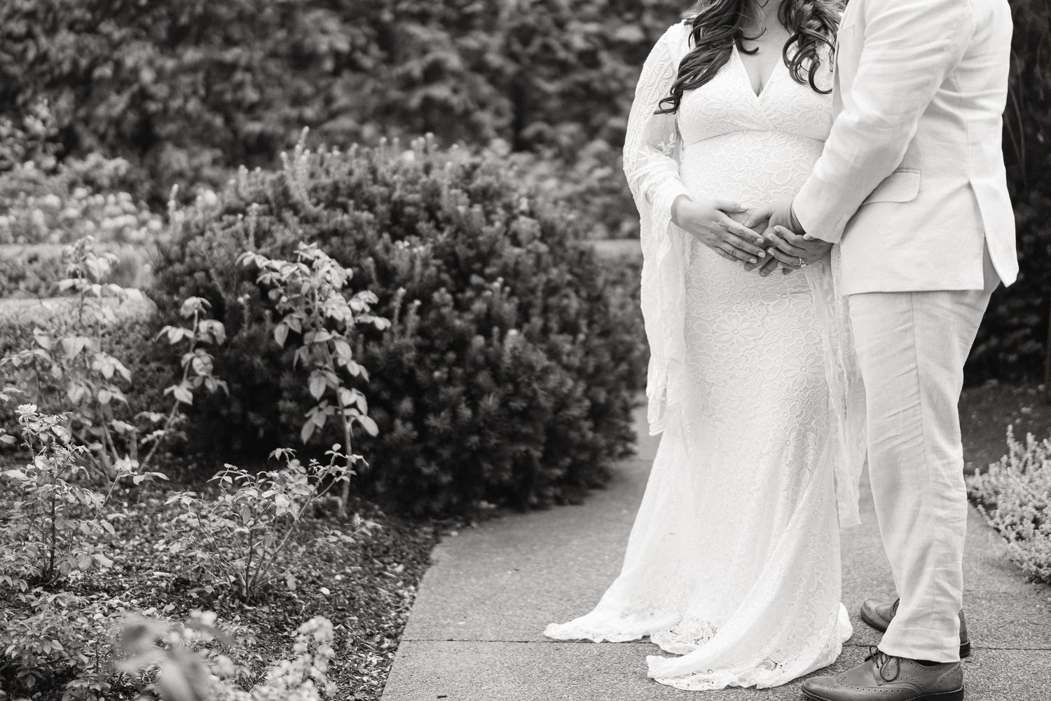 A couple in white wedding attire walks through a lush garden path during their outdoor wedding ceremony.