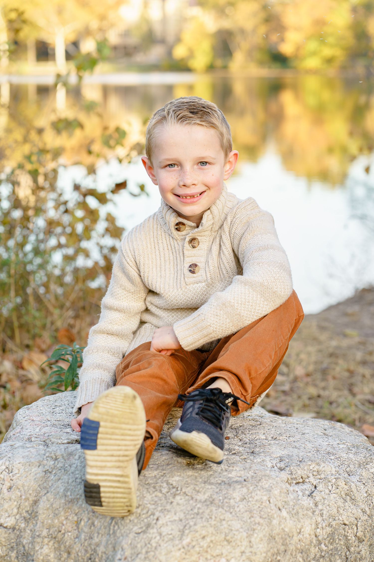 A child in a beige sweater and brown pants sits playfully on a large rock near a tranquil pond during autumn.