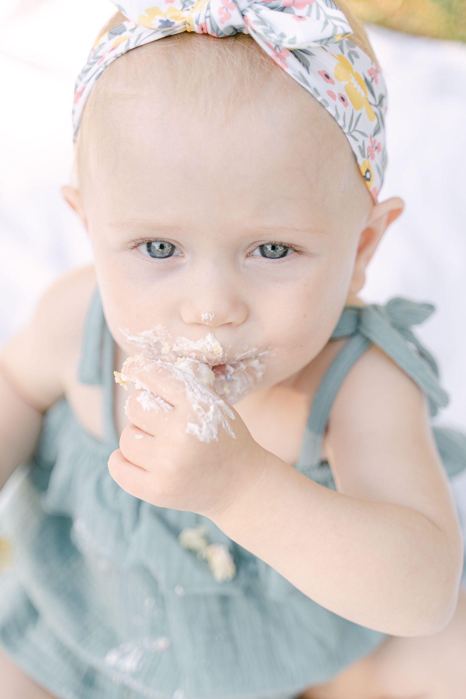 Baby wearing floral headband and mint dress enjoying cake smash photo session with frosting on face.