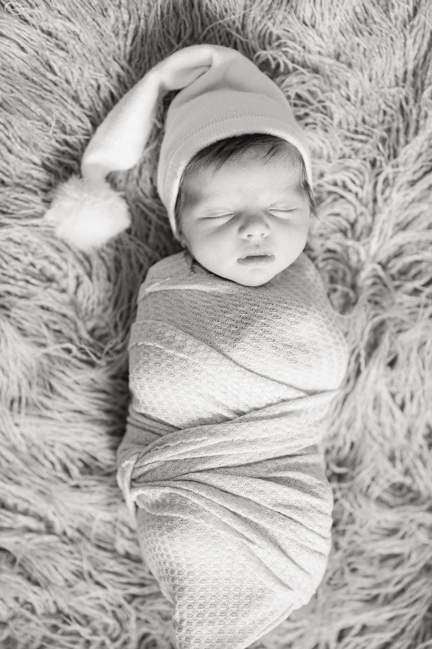 A sleeping newborn baby swaddled in white fabric lies on a soft fluffy gray blanket in black and white photography.