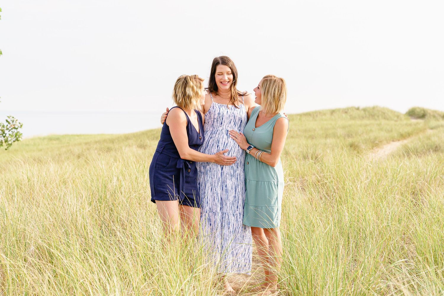 Three people wearing summer dresses stand together in a golden grass field at sunset.