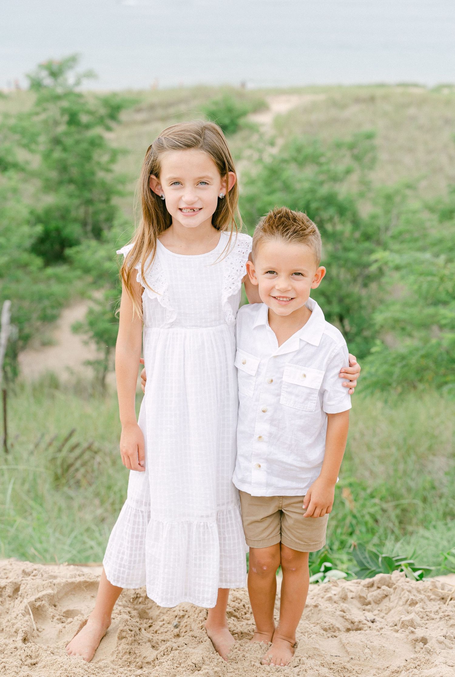 Two children in white outfits standing barefoot on sandy dune with natural backdrop.