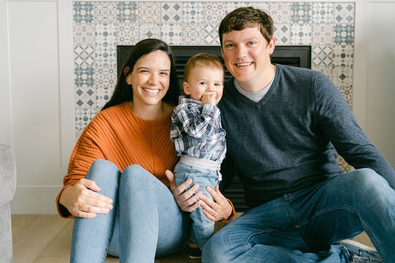 A family sits together on a grey couch in a living room with a patterned wallpaper background.