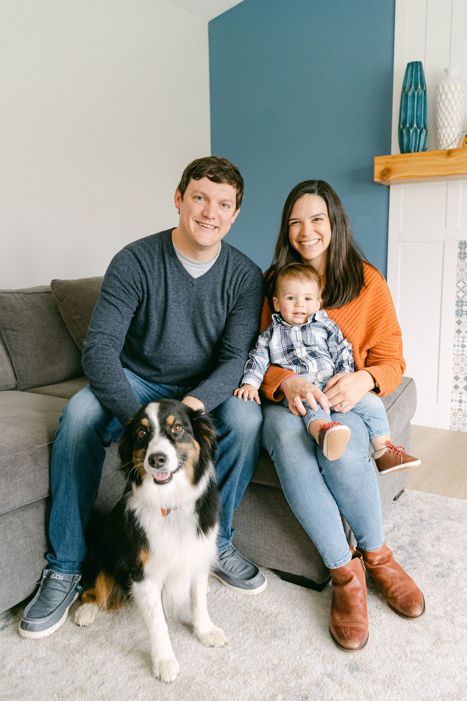 Family sits together on gray couch with their Australian Shepherd dog in living room with teal accent wall.