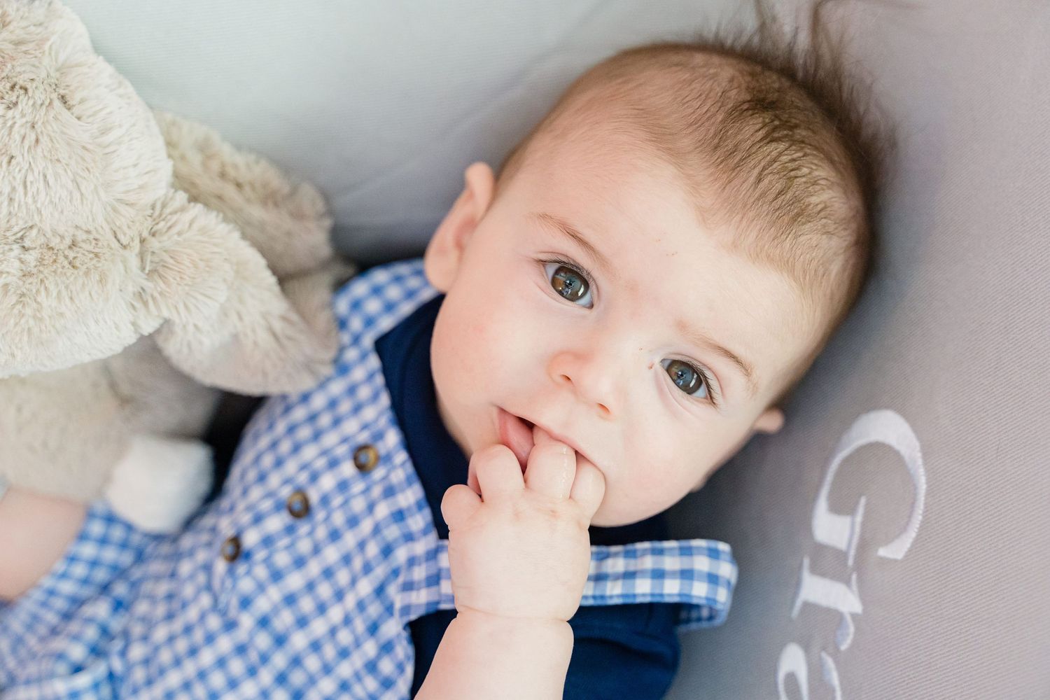 A baby wearing a blue checkered shirt lies on a white blanket while looking to the side.