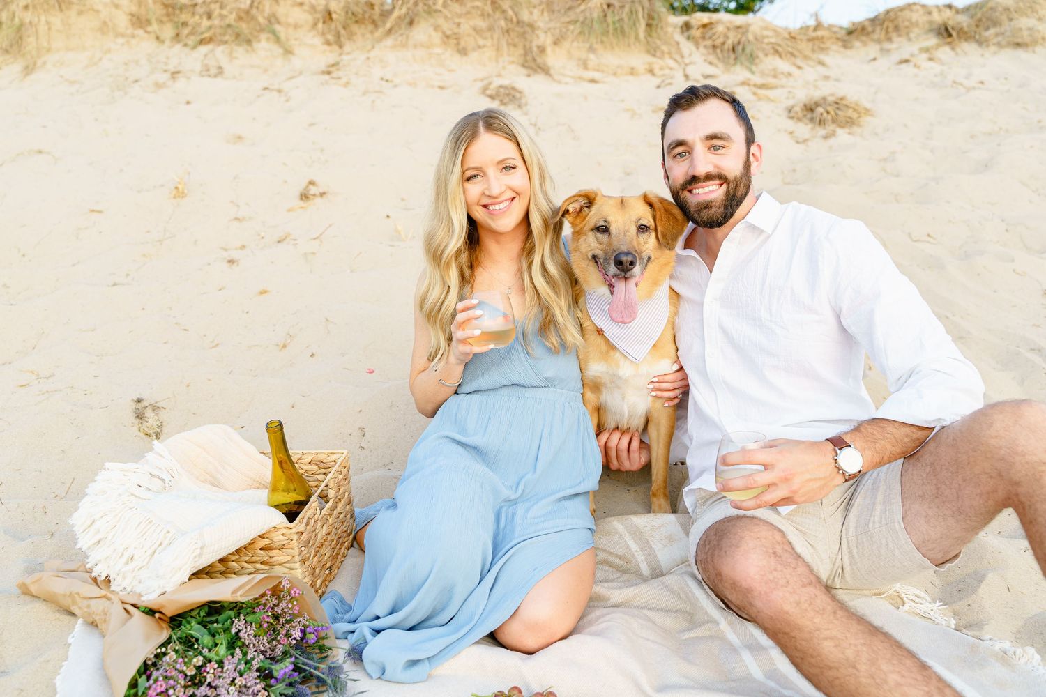 A happy couple sits on a sandy beach with their golden retriever dog enjoying a sunny afternoon together.